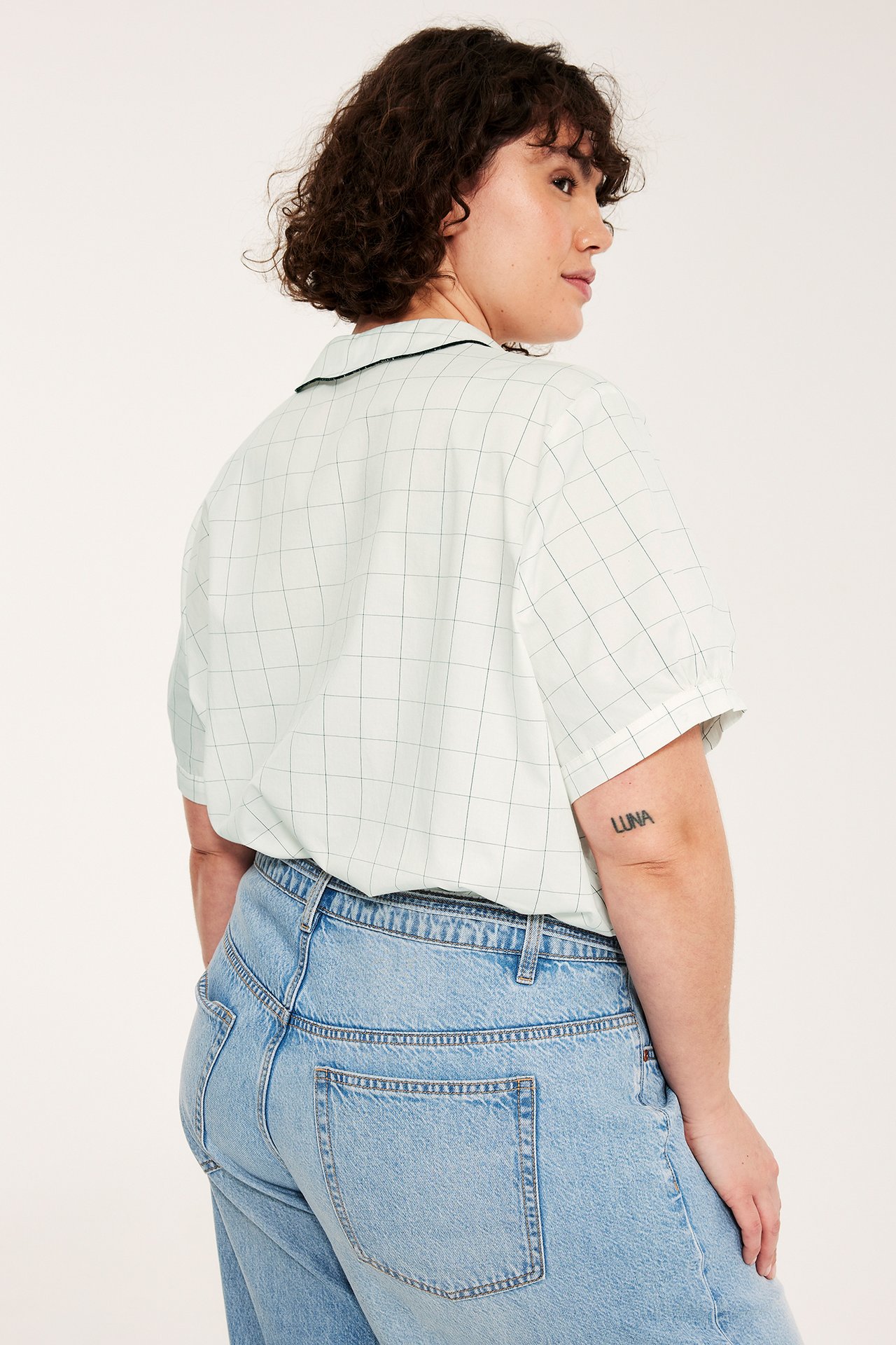 Bakifrån/sida: kvinna i offwhite rutig bomullsblus med vågig krage, svarta kanter, och ljusblå jeans.