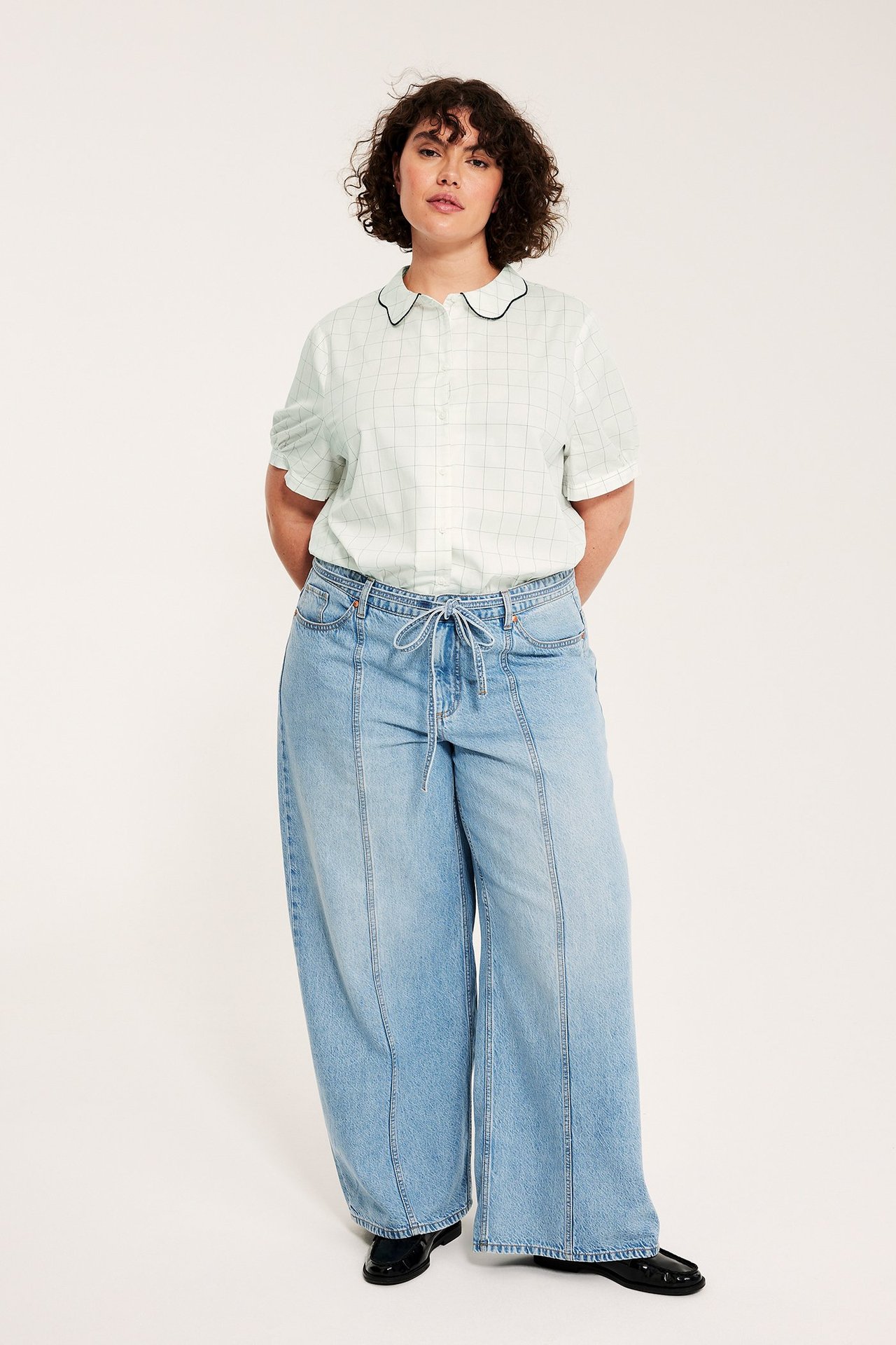 Nainen luonnonvalkoisessa ruudullisessa puuvillapuserossa mustalla aaltokauluksella, vaaleansiniset leveälahkeiset farkut, mustat kengät.