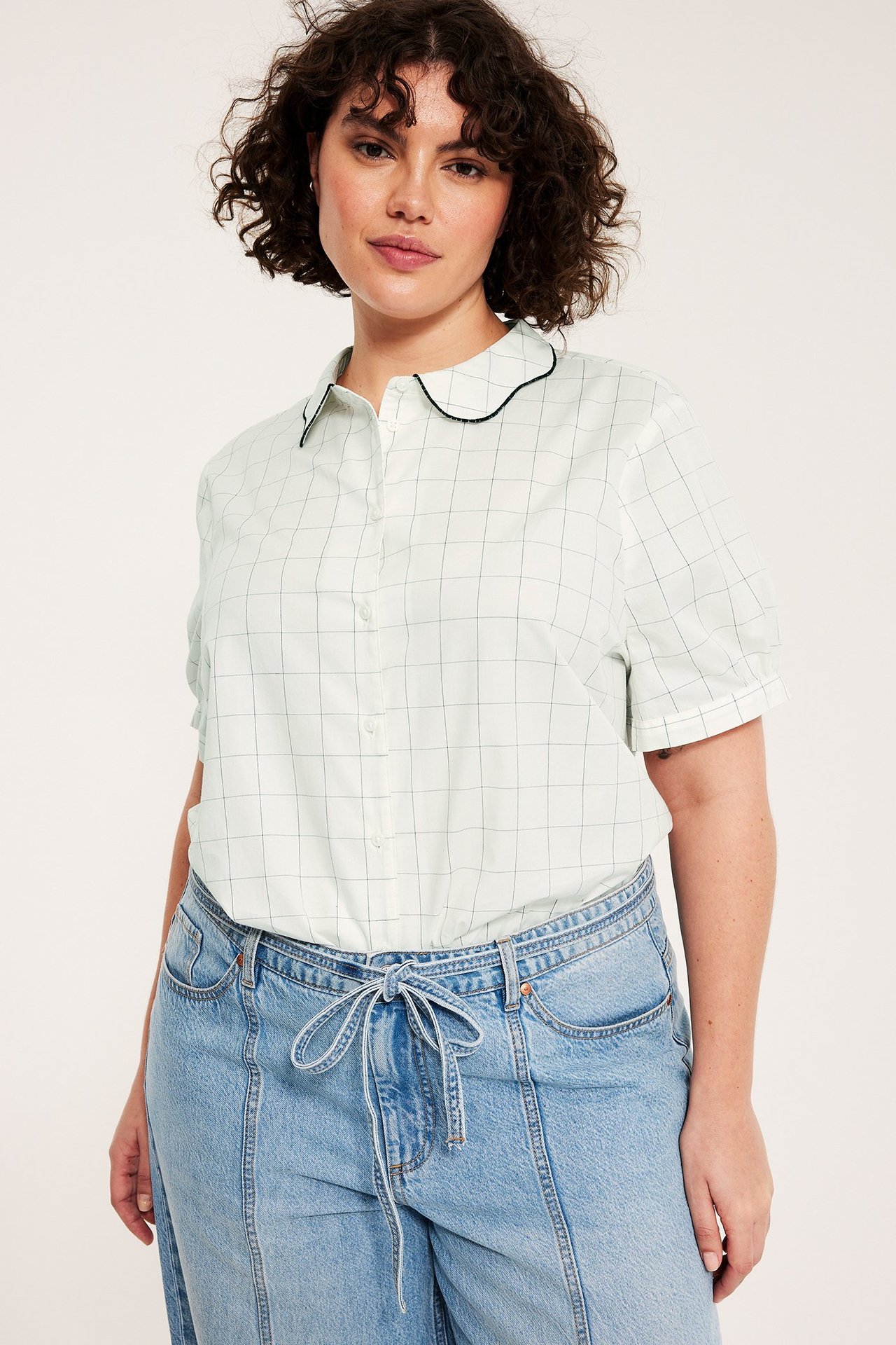 Edestä: Nainen luonnonvalkoisessa ruudullisessa puuvillapuserossa, mustareunainen aaltokaulus ja vaaleansiniset farkut.