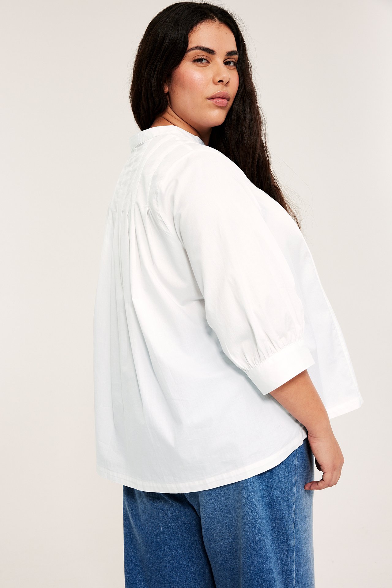 Sned bakifrån-vy av kvinna i en vit kraglös bomullsblus med 3/4 puffärm och blå vida jeans.