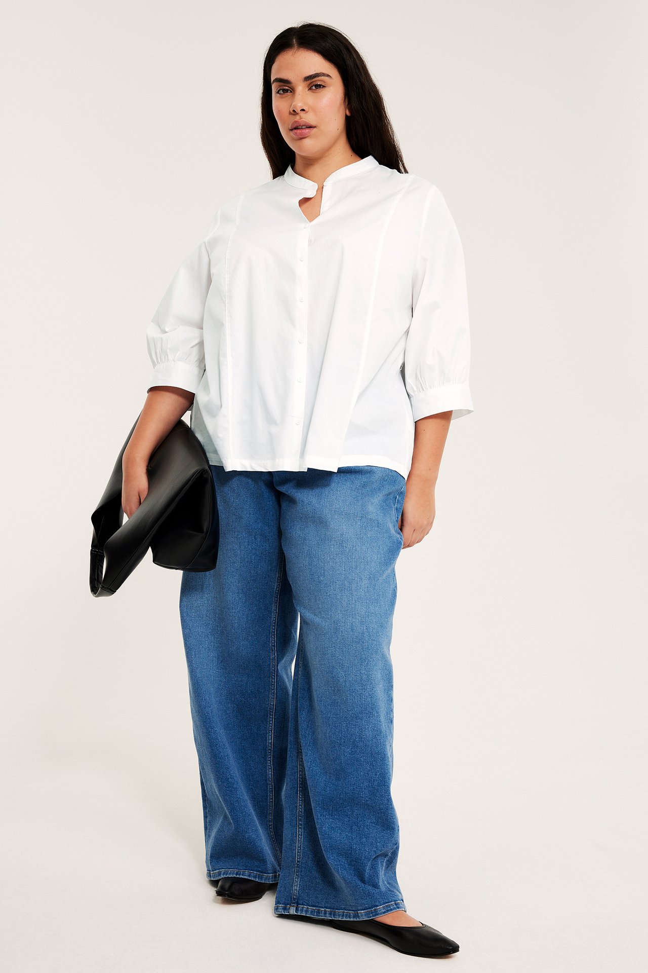 Widok z przodu: kobieta w białej bawełnianej bluzce z bufiastymi rękawami, niebieskich szerokich jeansach, czarnych butach, trzyma torebkę.