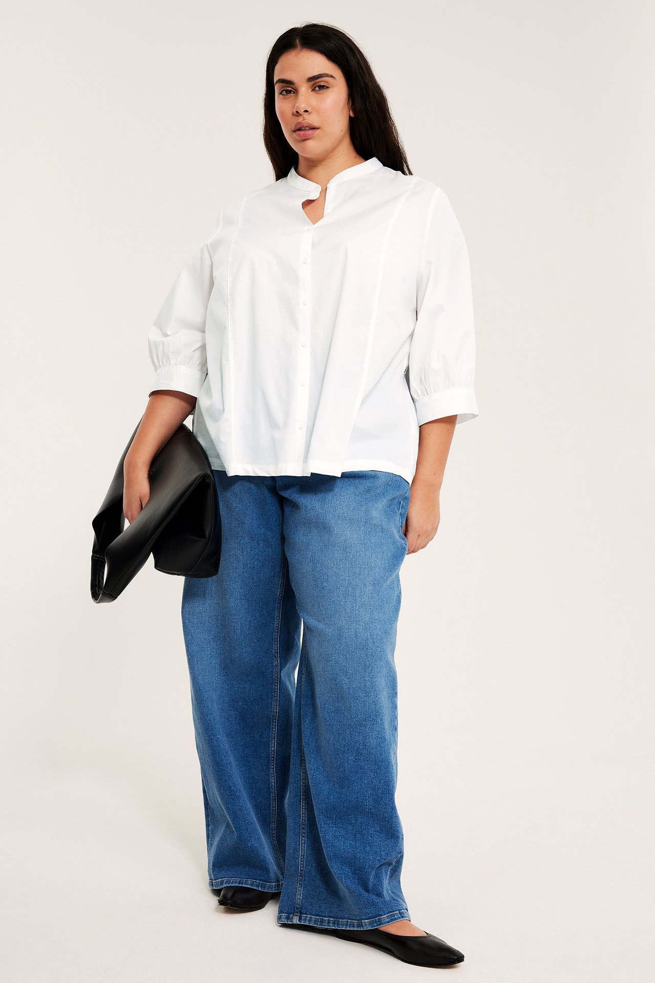 Widok z przodu: kobieta w białej bawełnianej bluzce z bufiastymi rękawami, niebieskich szerokich jeansach, czarnych butach, trzyma torebkę.