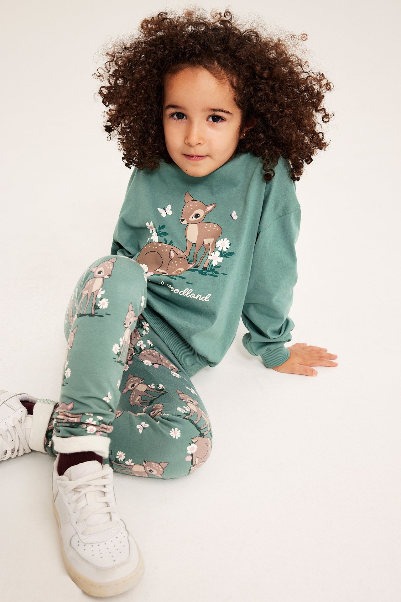 Edestä: Lapsi vihreissä kuviollisissa leggingseissä, joissa peura- ja kukkaprintti, sekä vastaava vihreä collegepaita.