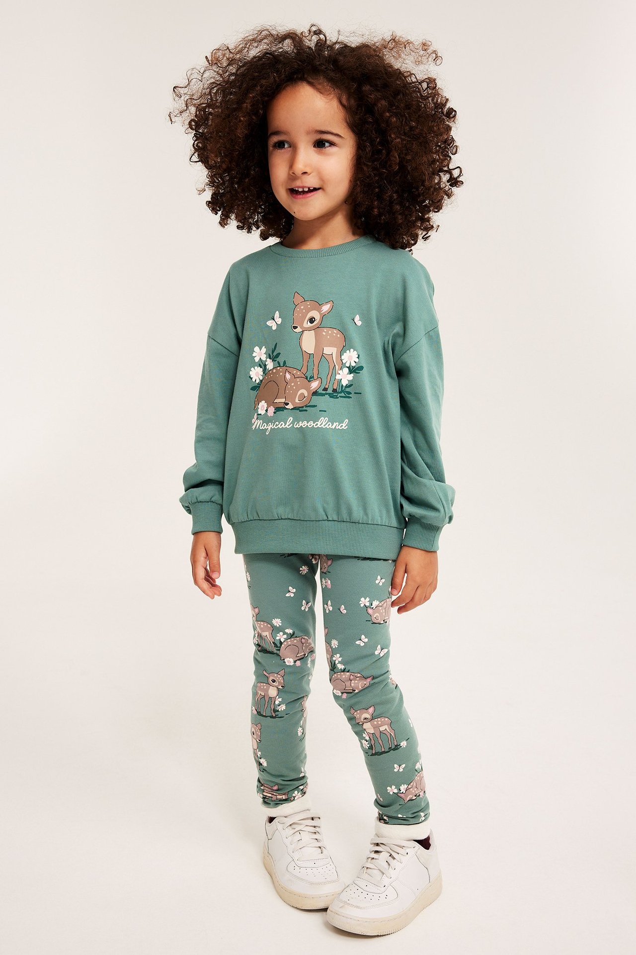 Widok z przodu: dziecko w zielonych wzorzystych legginsach w sarny i kwiaty, pasująca bluza i białe trampki.
