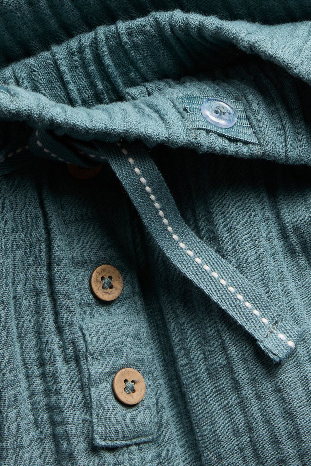 Nærbilde av mørketurkis bomullsmuslinbukse med elastisk midje, fiskebeinssnor og dekorative treknapper.