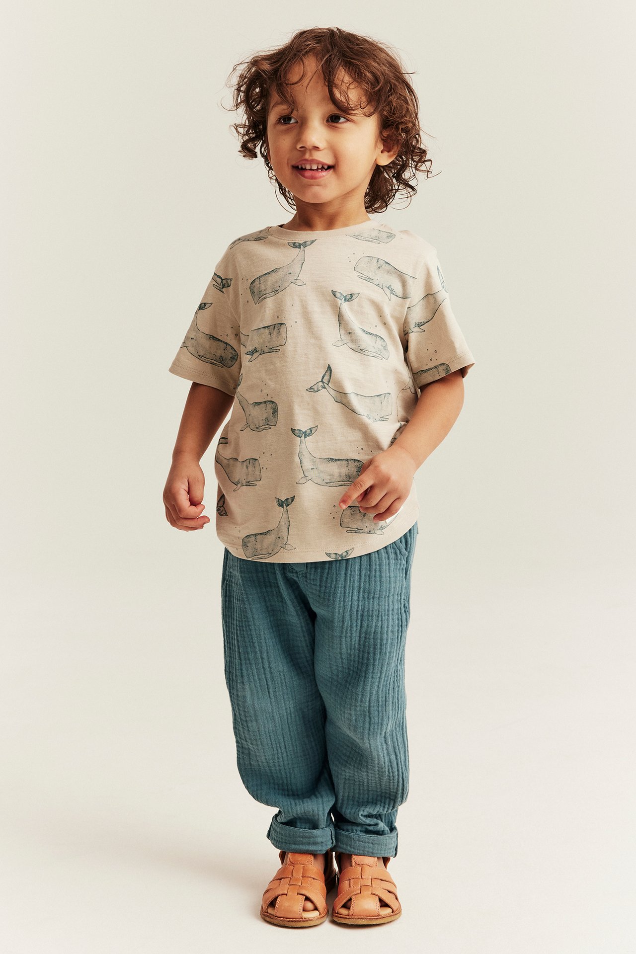 Widok: Dziecko nosi ciemnoturkusowe muślinowe spodnie, beżowy t-shirt z nadrukiem wielorybów i brązowe sandały.