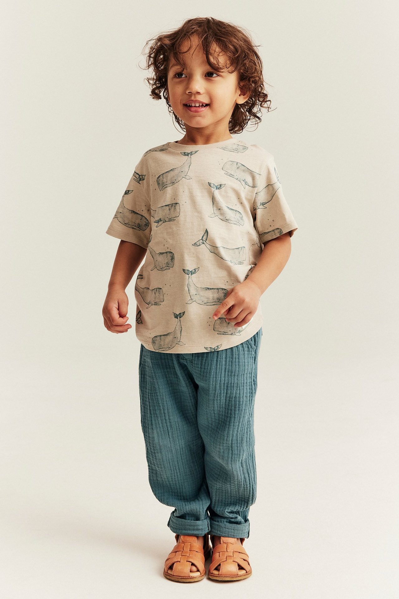 Widok: Dziecko nosi ciemnoturkusowe muślinowe spodnie, beżowy t-shirt z nadrukiem wielorybów i brązowe sandały.