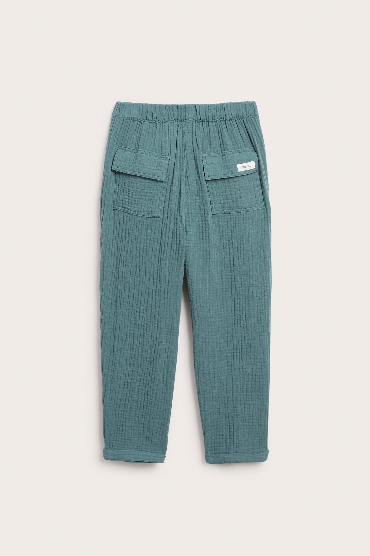 Bakfra: mørk turkis bomullsmuslinbukse med elastisk midje, to baklommer med klaff og opprullede benkanter.
