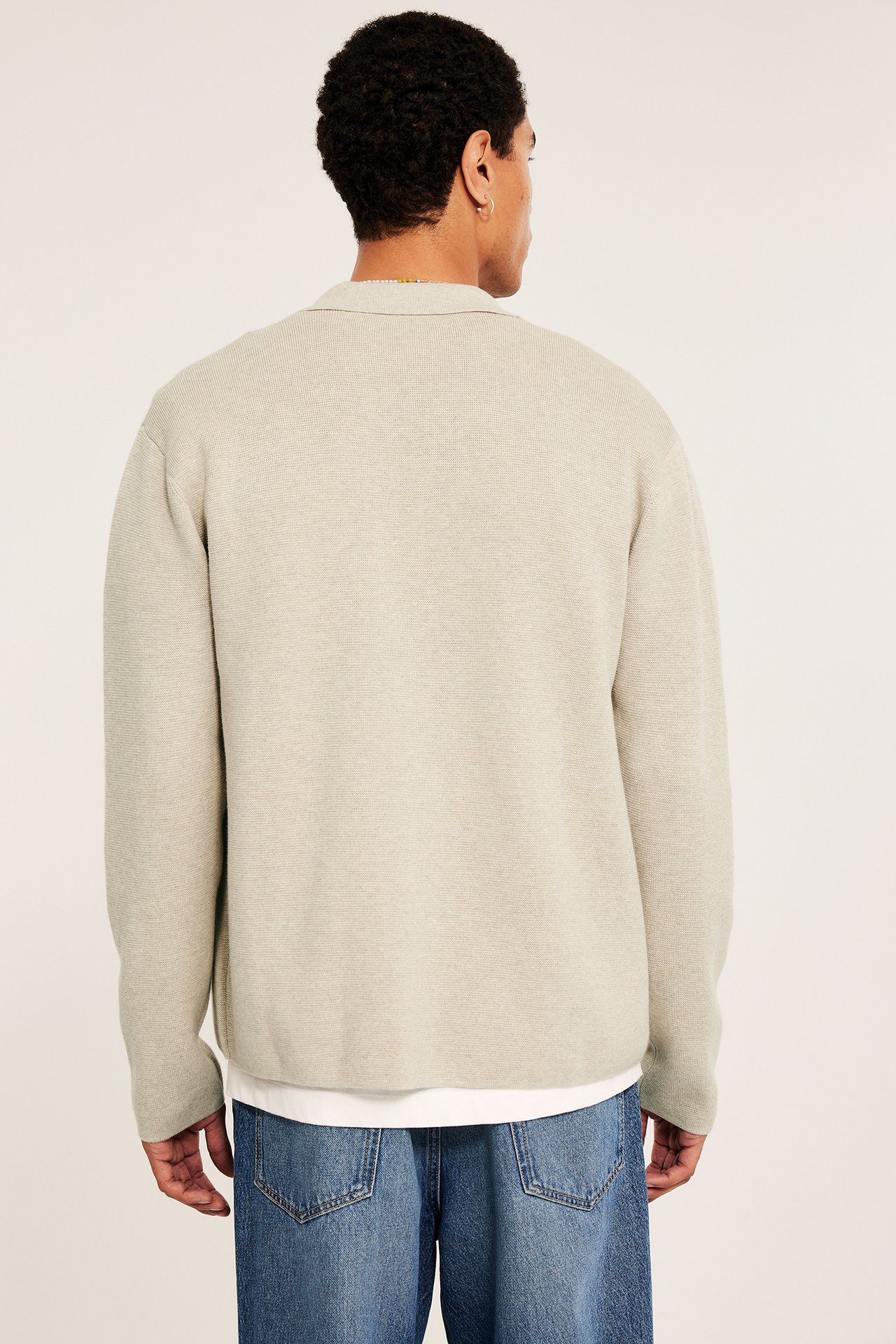 Bakfra av en mann i en beige melert strikket kragecardigan over en hvit t-skjorte og blå jeans.