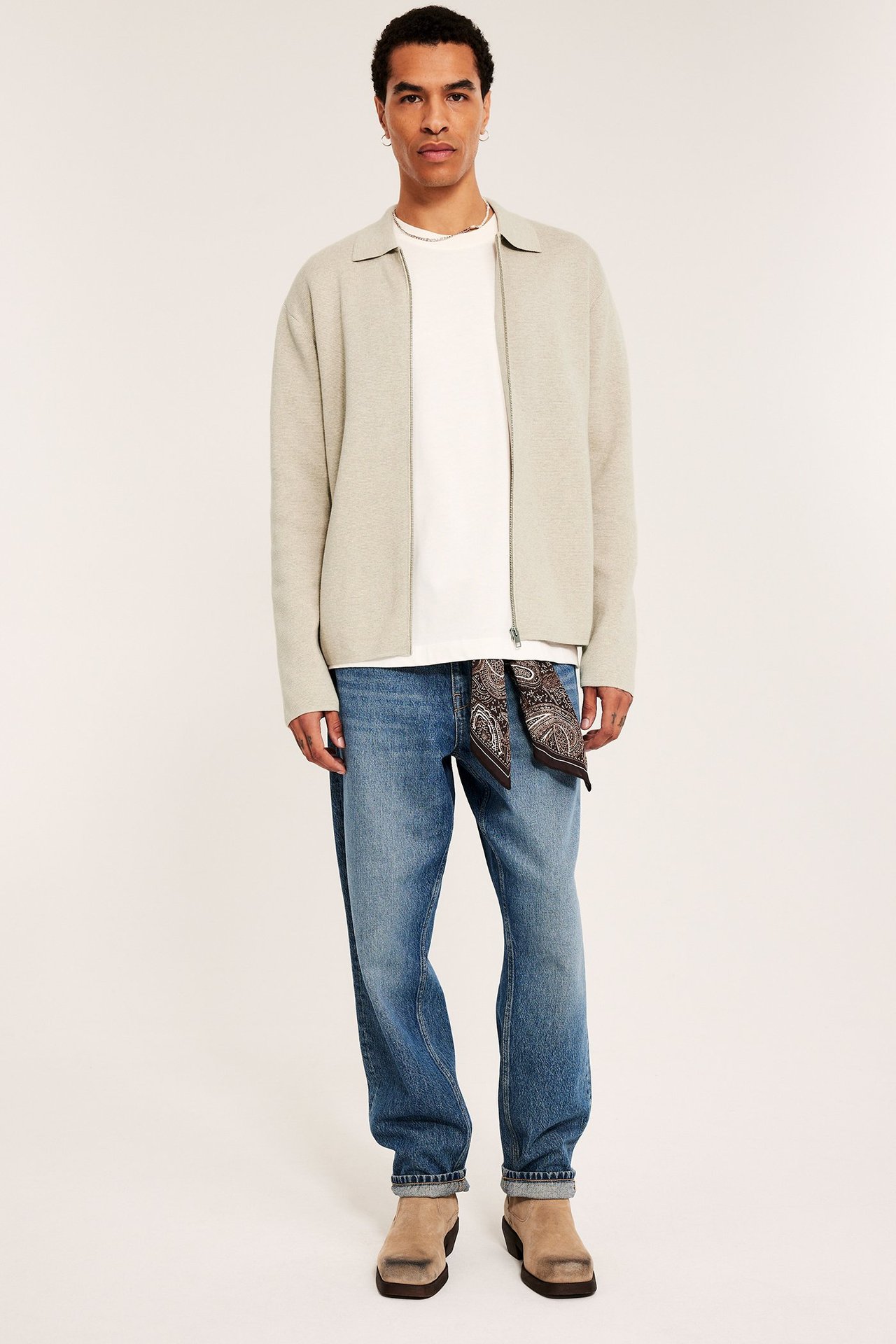 Mann i beigemelert strikket cardigan, hvit t-skjorte, blå jeans og beige boots.