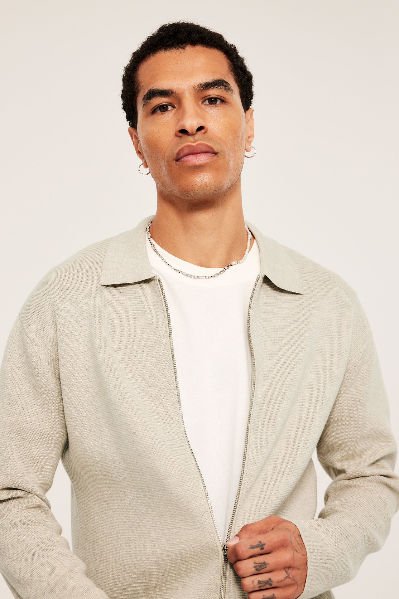 Widok z przodu: mężczyzna zapinający beżowy melanżowy bawełniany kardigan na białą koszulkę, ze srebrnymi naszyjnikami.