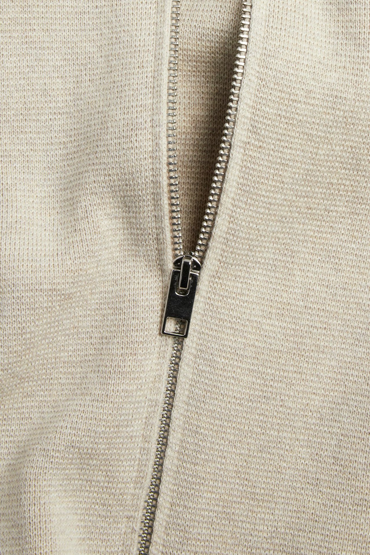 Nærbilde av en beige melert strikket bomullscardigan med sølvfarget glidelås foran.