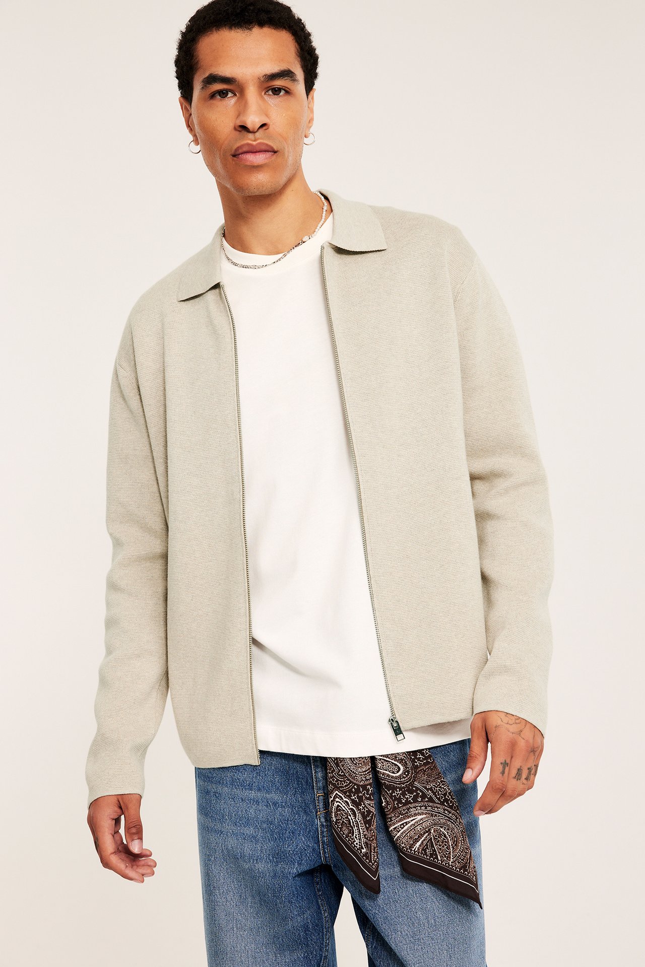 Forfra: Mann har på seg åpen melert beige strikket cardigan, hvit t-skjorte, blå jeans og bandana.
