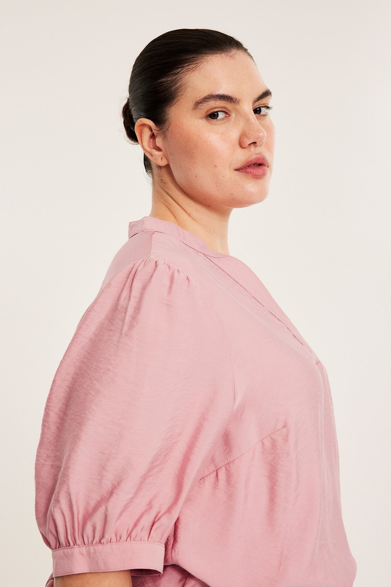 Sidebilde av en kvinne i rosa pop-over-bluse med V-hals og 3/4 puffermer.