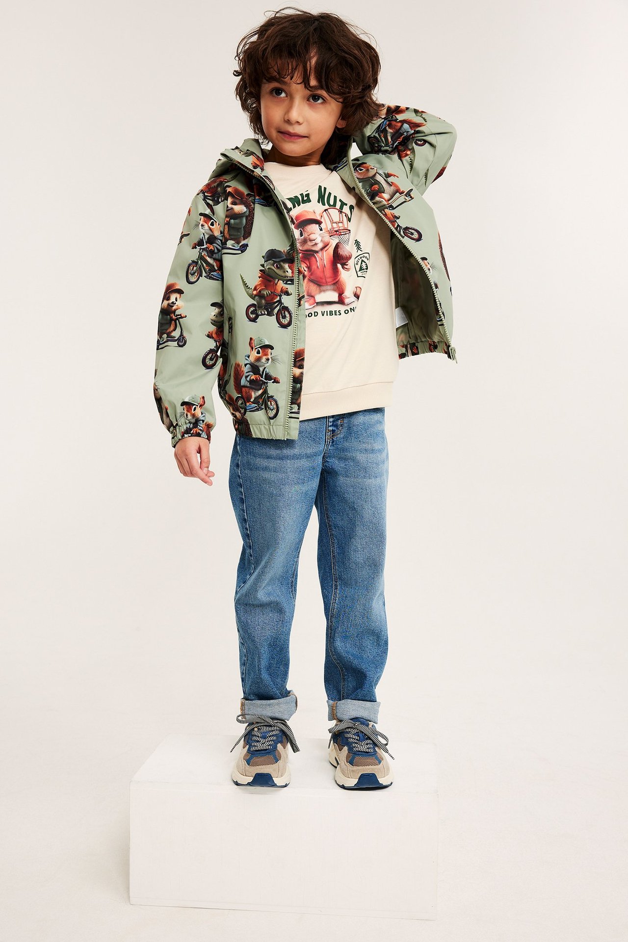 Lapsi tummanvihreässä kuviollisessa takissa, vaaleanbeigessä collegepaidassa, sinisissä farkuissa ja tennareissa, edestä.