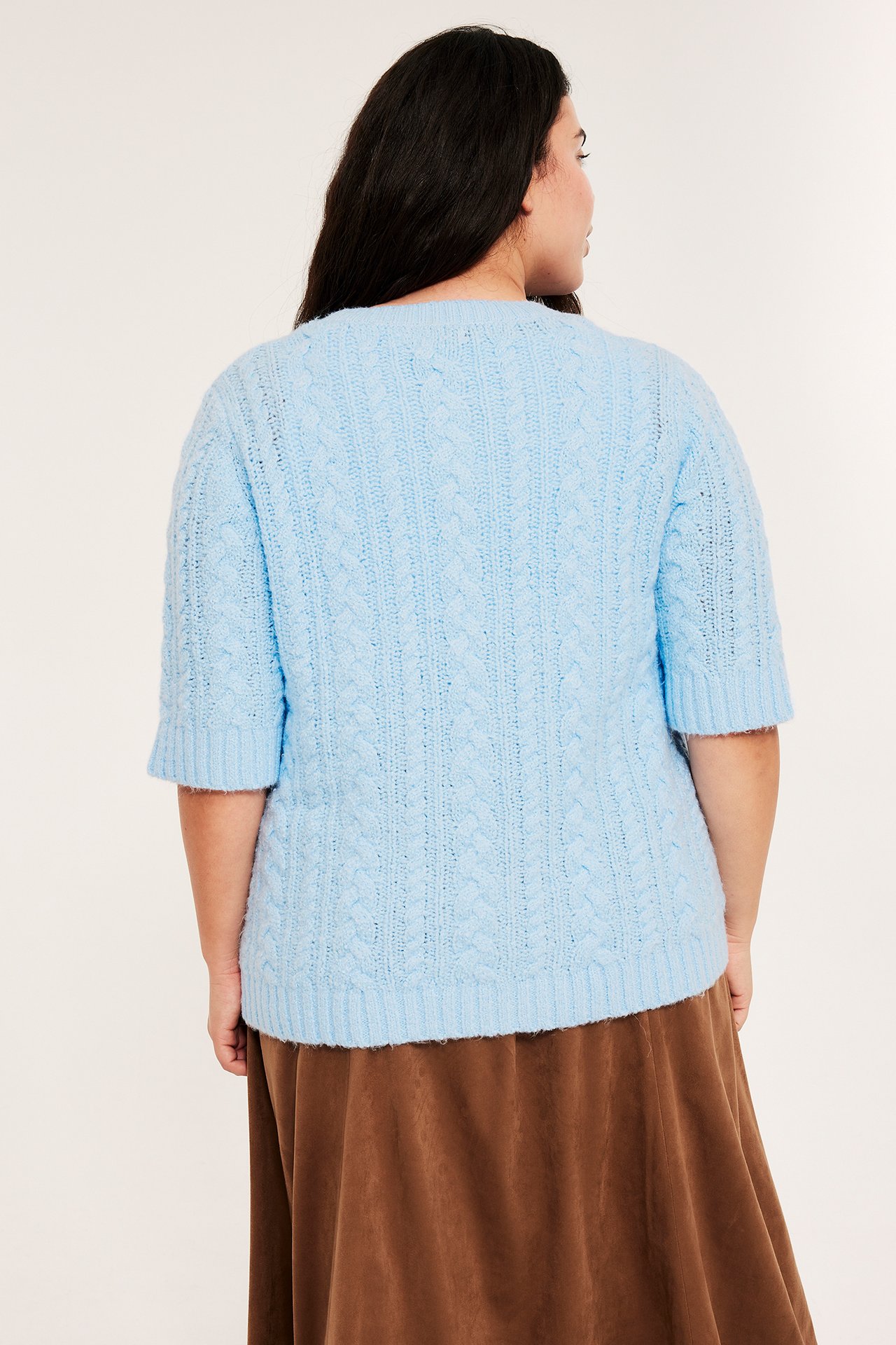 Bakfra: En kvinne i lys blå kabelstrikket kortermet cardigan over et brunt skjørt.