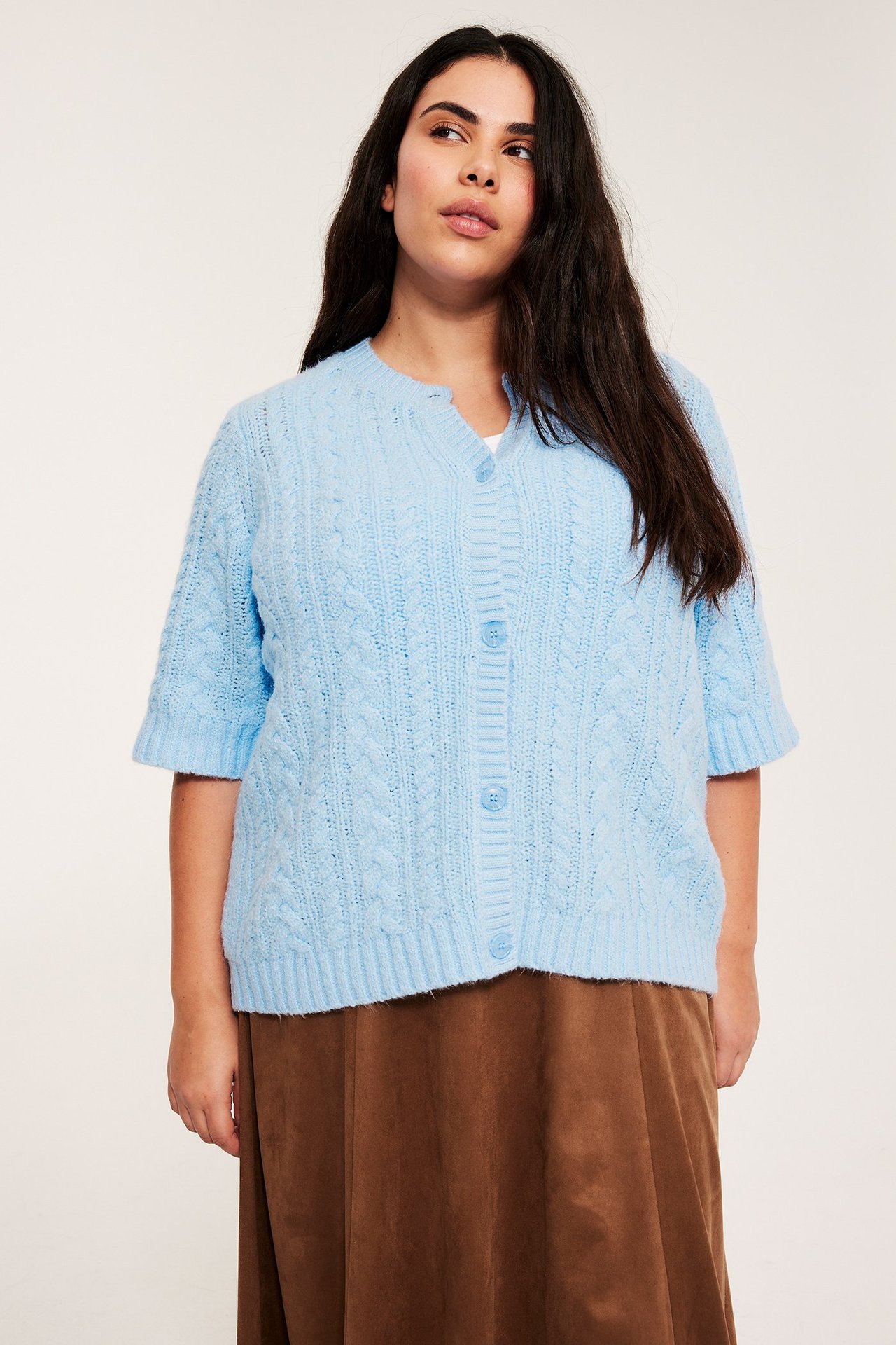 Framifrån: kvinna i ljusblå kabelstickad kortärmad kofta med knappar, över en brun kjol.