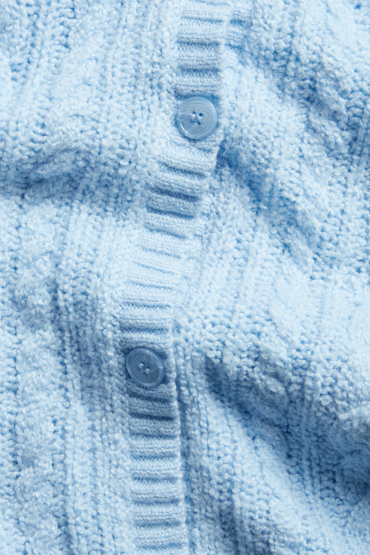 Nærbilde av en lyseblå kabelstrikket cardigan med to lyseblå knapper foran.