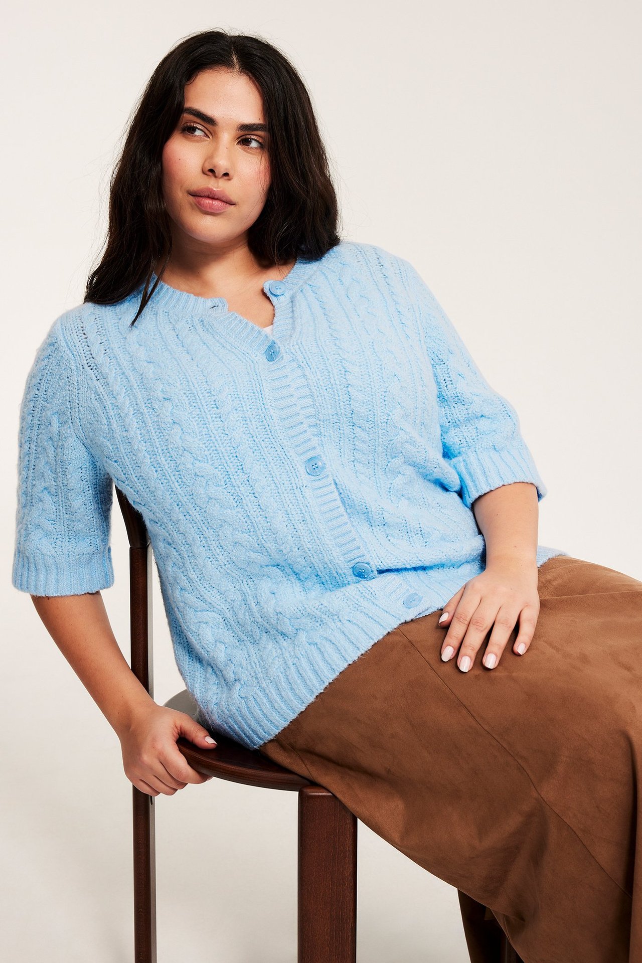 Frontbild på en kvinna i ljusblå kabelstickad kortärmad kofta med knappar och brun kjol.