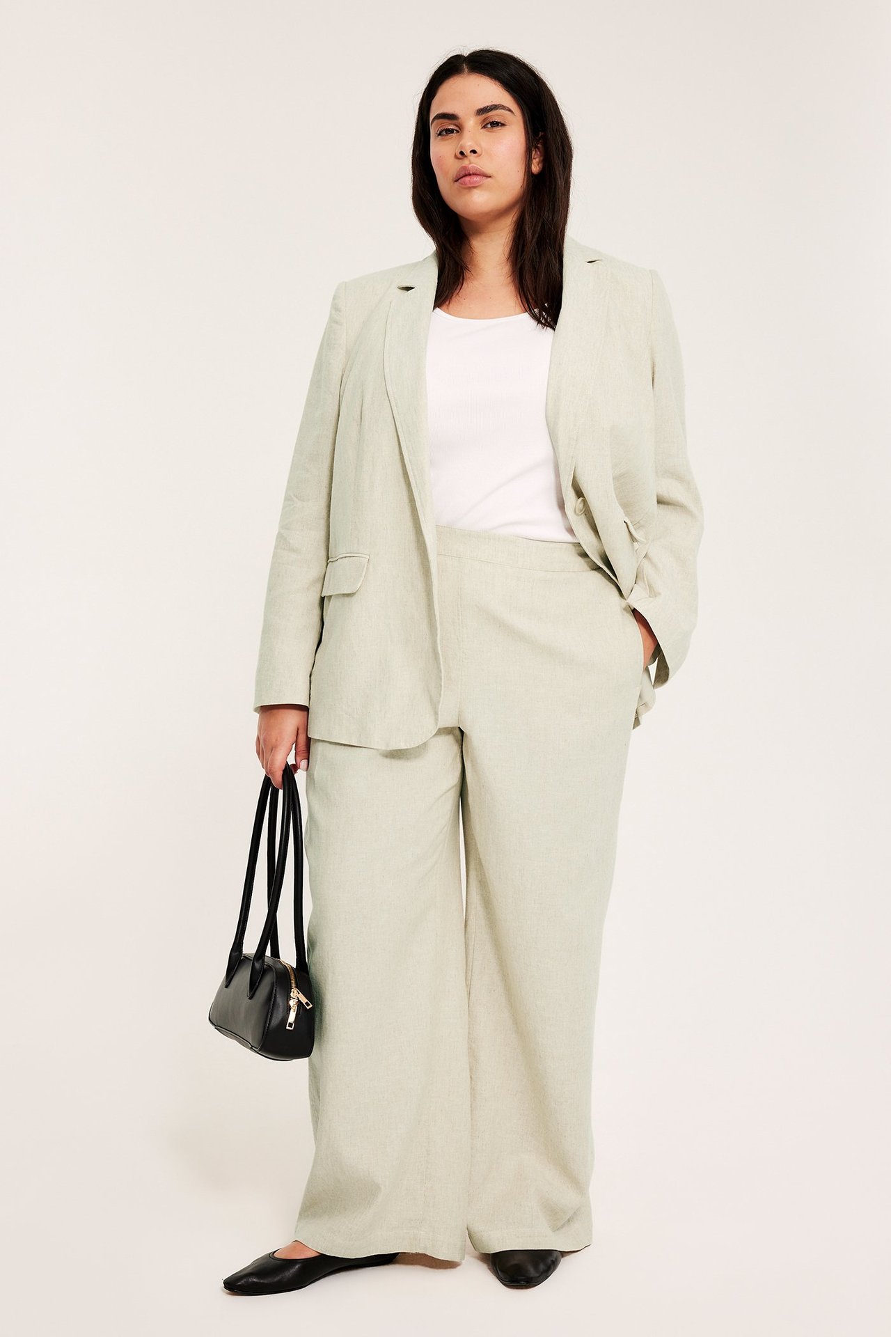 Edestä: Nainen beigessä pellavasekoitekavajassa, yhteensopivissa housuissa, valkoisessa t-paidassa, musta käsilaukku ja ballerinat.