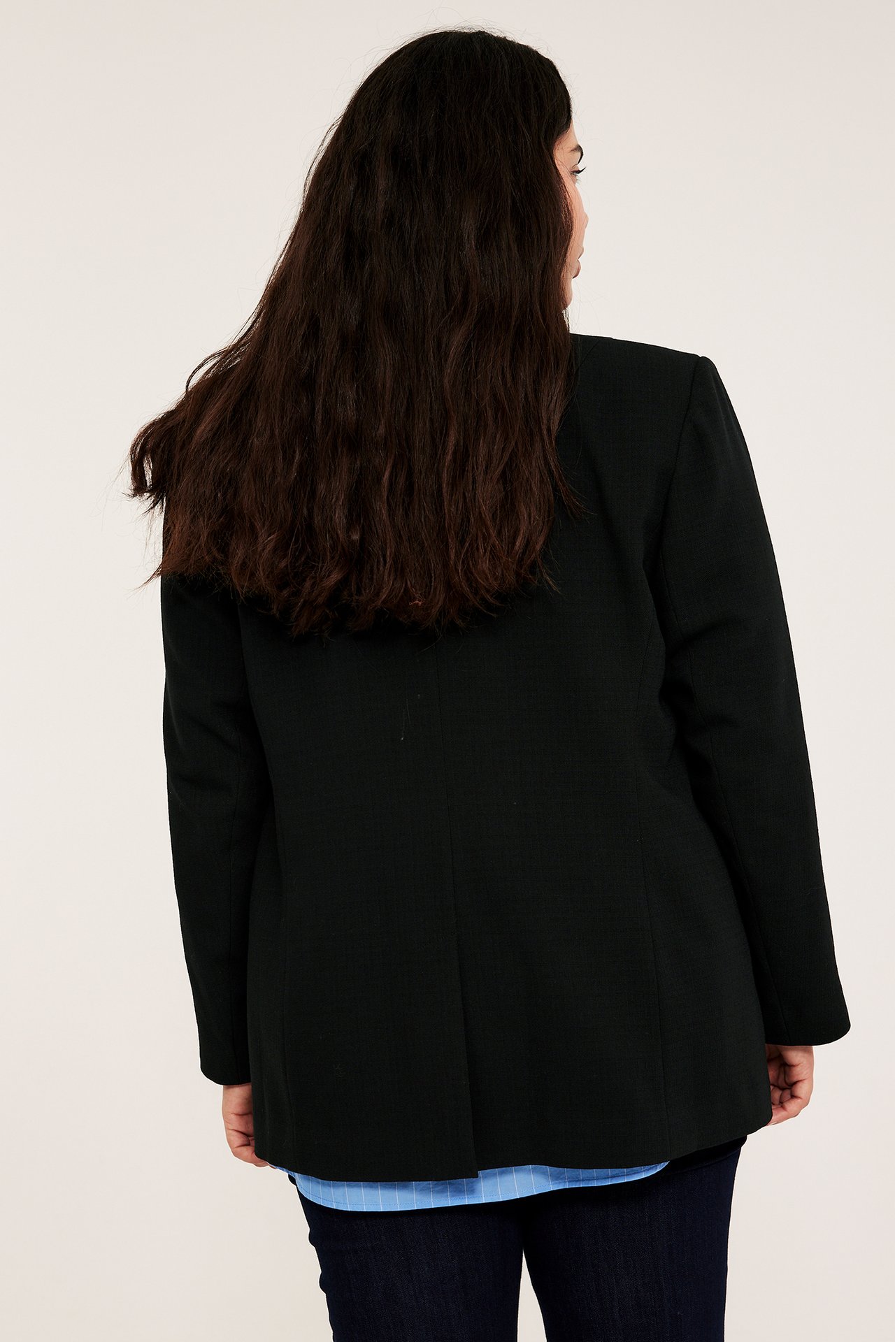 Naisen takakuva mustassa strukturoidussa bleiserissä, alla siniraitainen paita ja tummansiniset farkut.