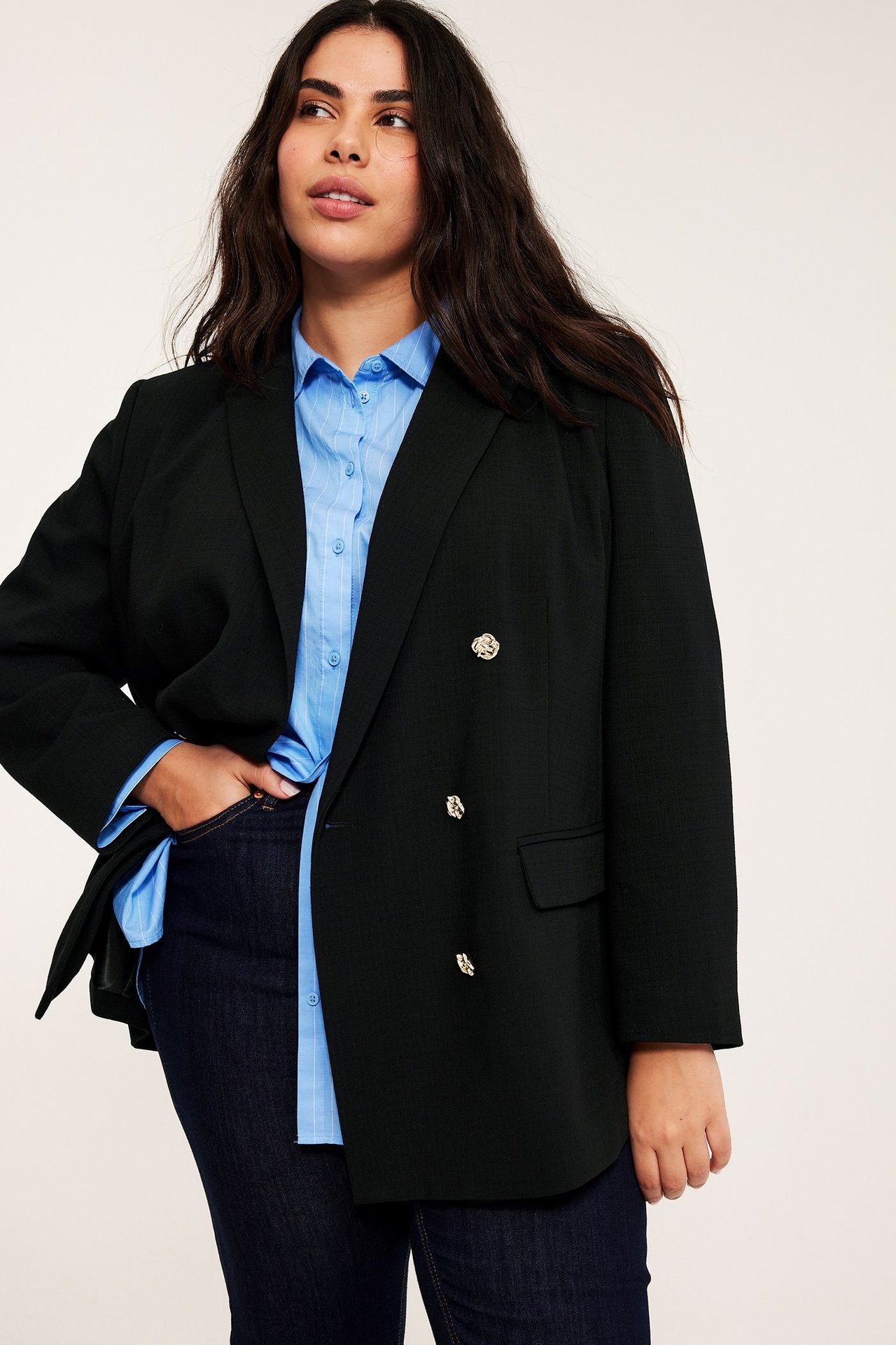 Etunäkymä: Nainen mustassa kaksirivisessä bleiserissä kultanapeilla, vaaleansinisessä raidallisessa paidassa, tummissa farkuissa.