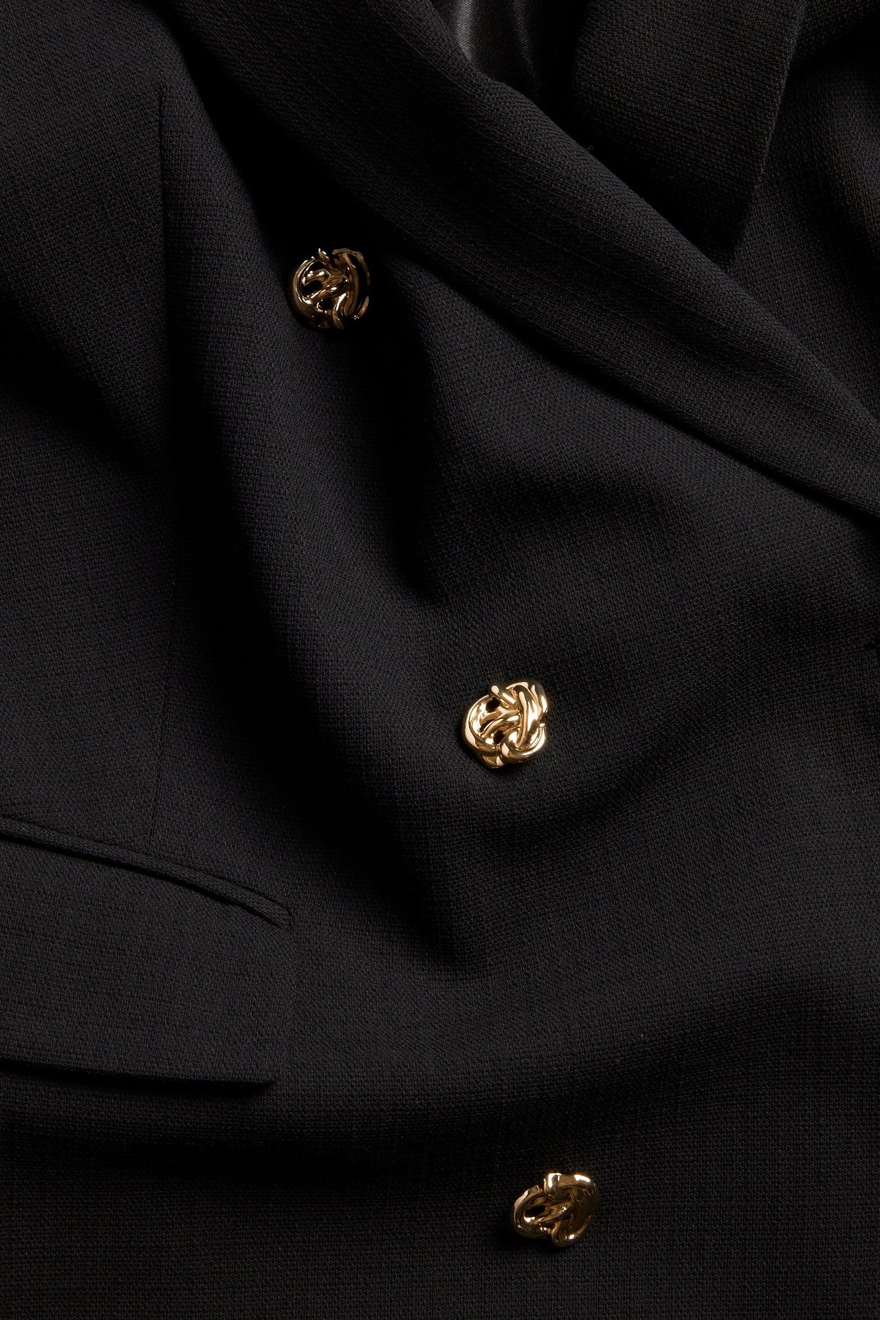 Zbliżenie czarnej, dwurzędowej marynarki z tkaniny strukturalnej z trzema ozdobnymi złotymi guzikami i detalem kieszeni.