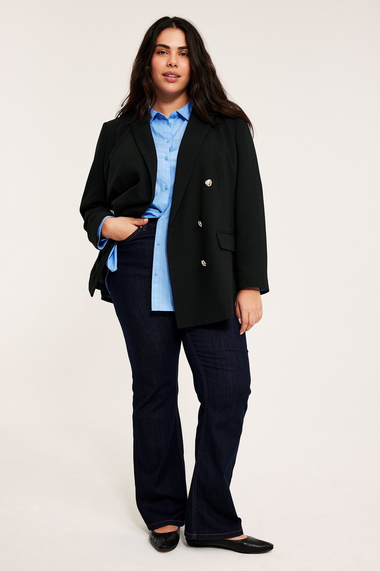 Kobieta od przodu: w czarnej dwurzędowej marynarce ze złotymi guzikami, jasnoniebieskiej koszuli i ciemnych dżinsach.