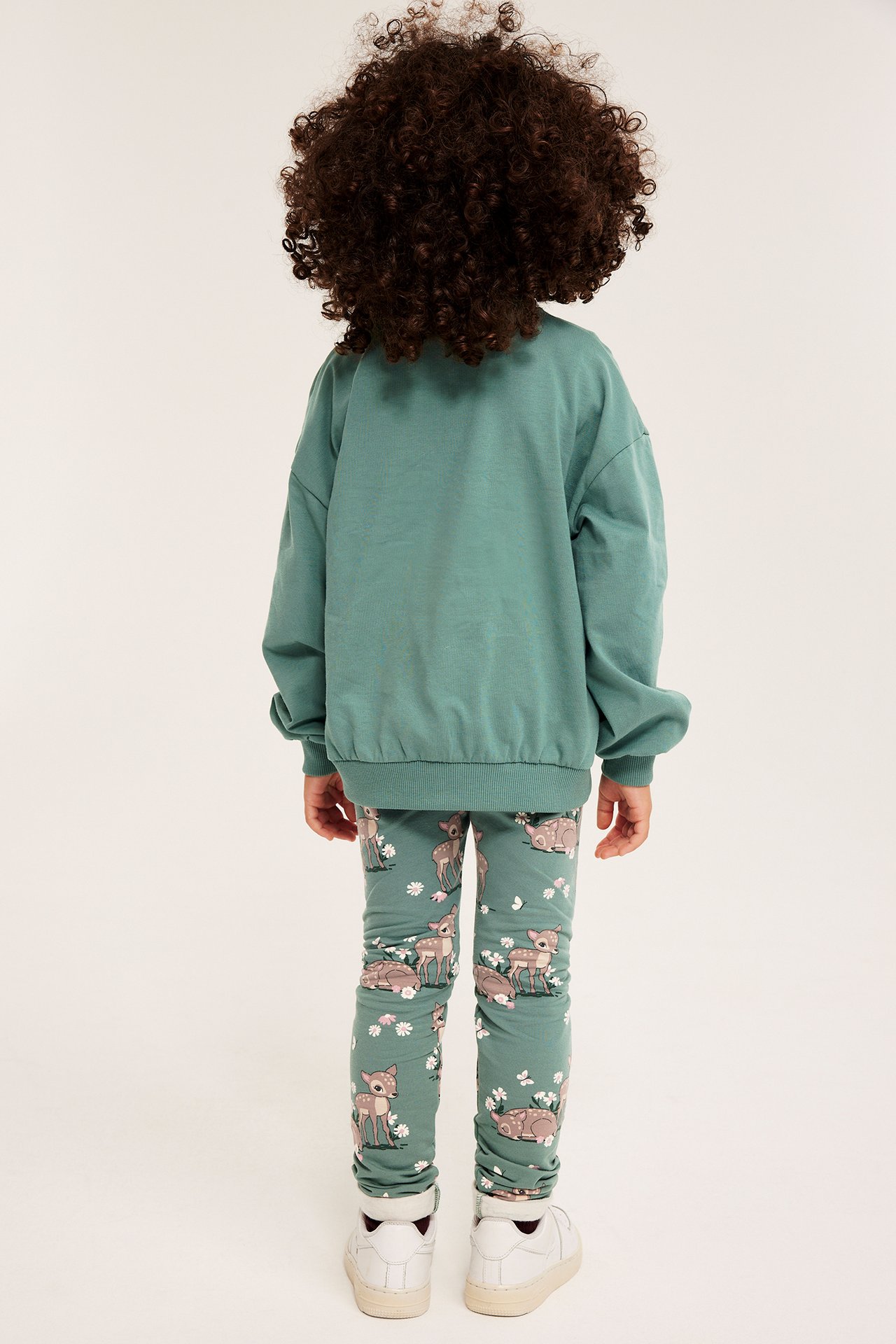 Ryggbilde av et barn iført grønn bomullssweatshirt og grønne leggings med rådyr- og blomstermønster.