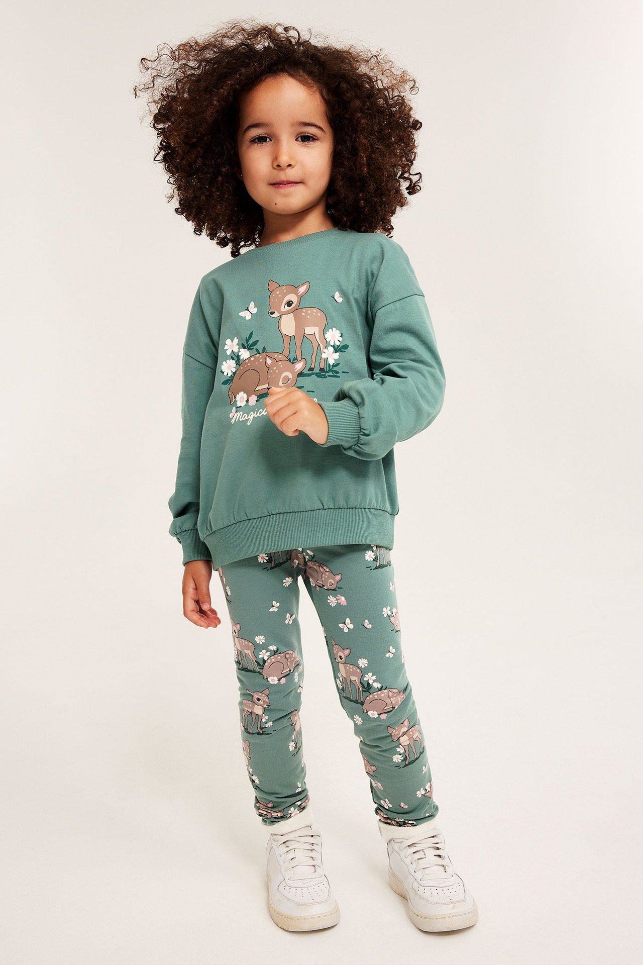 Edestä kuvattuna lapsi vihreässä puuvillapaidassa, jossa peuroja, ja yhteensopivissa trikoissa.