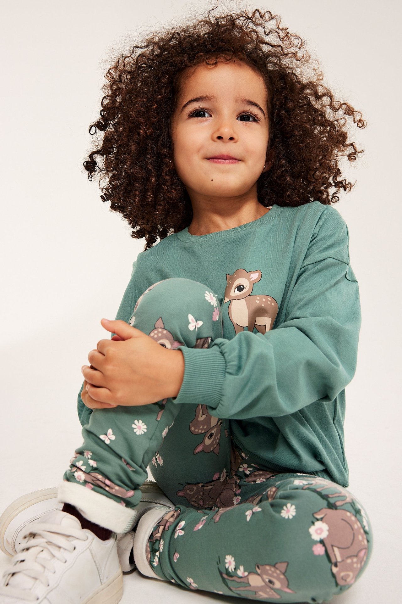 Z przodu, dziecko w zielonej bawełnianej bluzie z nadrukiem sarenki, pasujących zielonych dresach i białych trampkach.