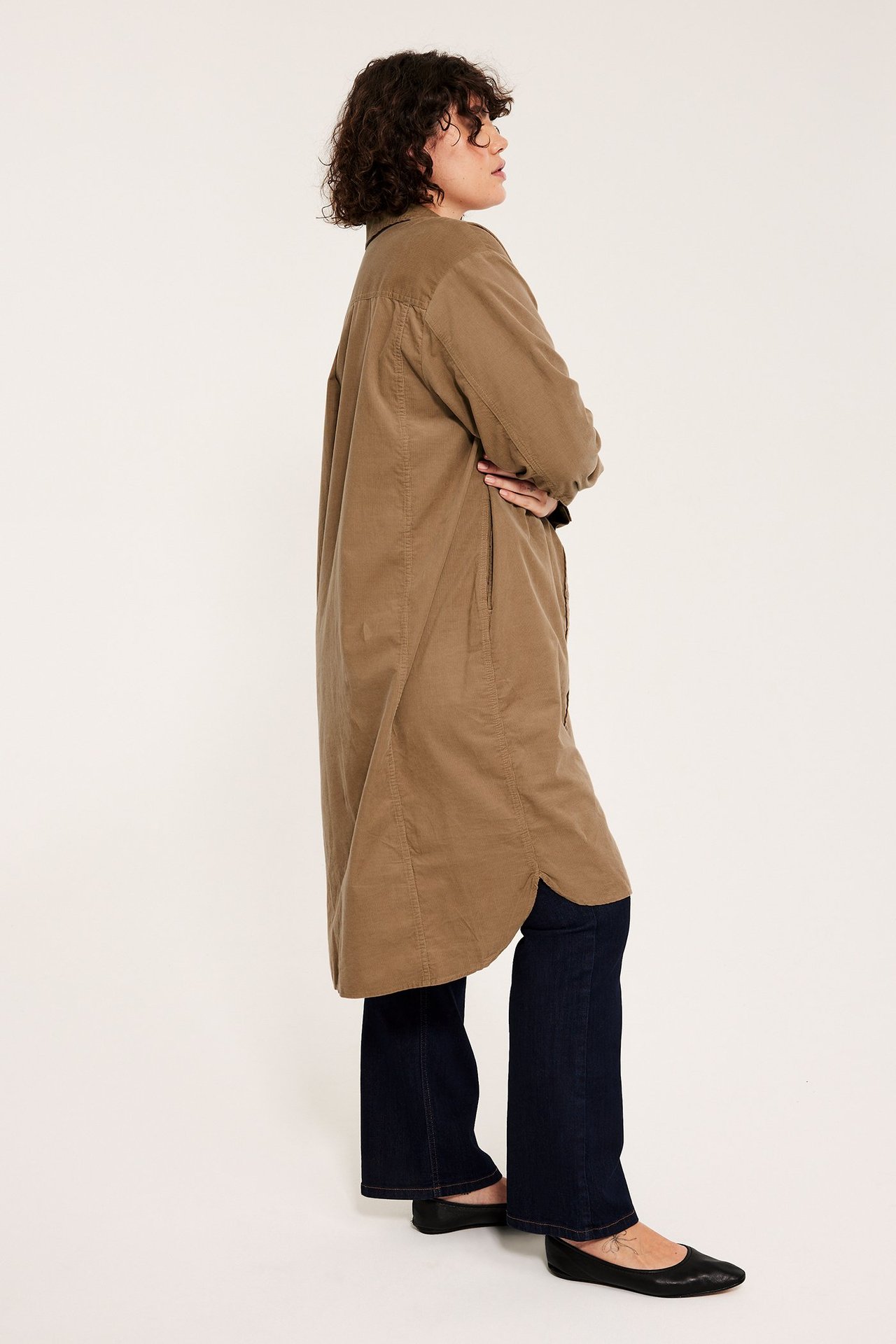 Sideprofil av en kvinne i en kalvelang brun cordfløyels-skjortekjole, mørkeblå bukser og svarte flate sko.