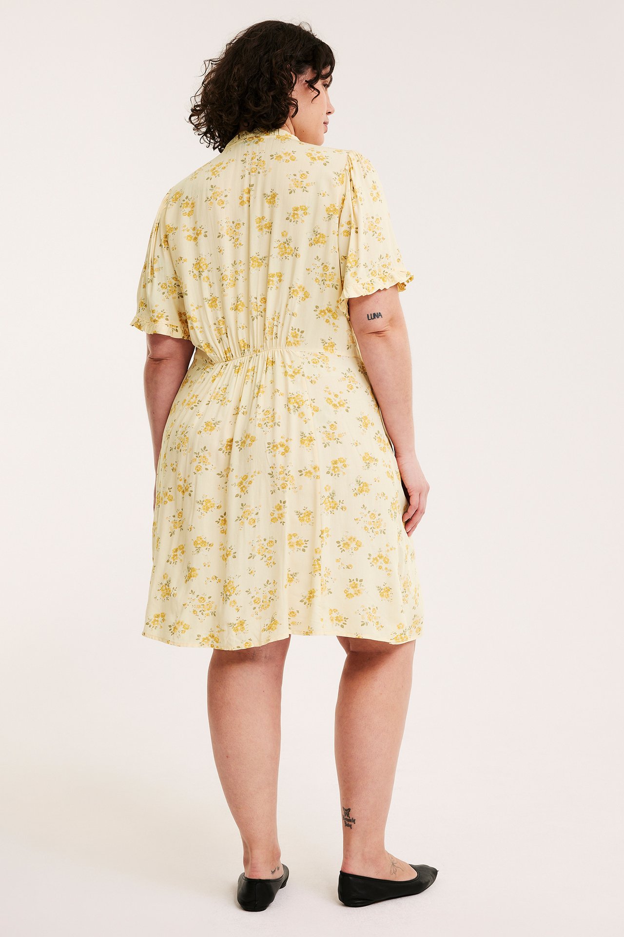 Ryggvy av en kvinna i en ljusgul blommig viskosklänning med rynkad midja och volangärmar.