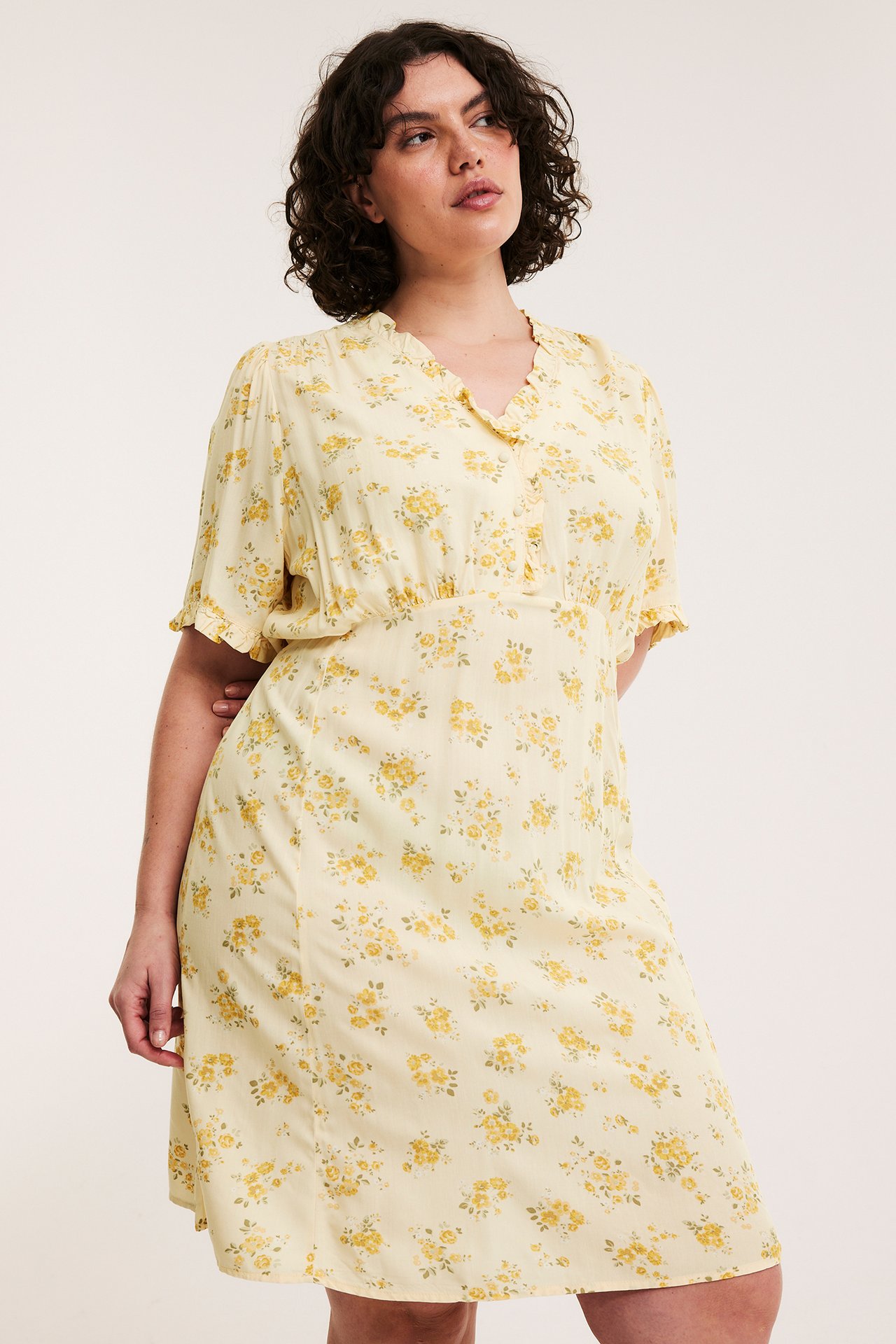 Framsida: kvinna i ljusgul blommig viskosklänning, knälång, med volang V-hals och korta ärmar.