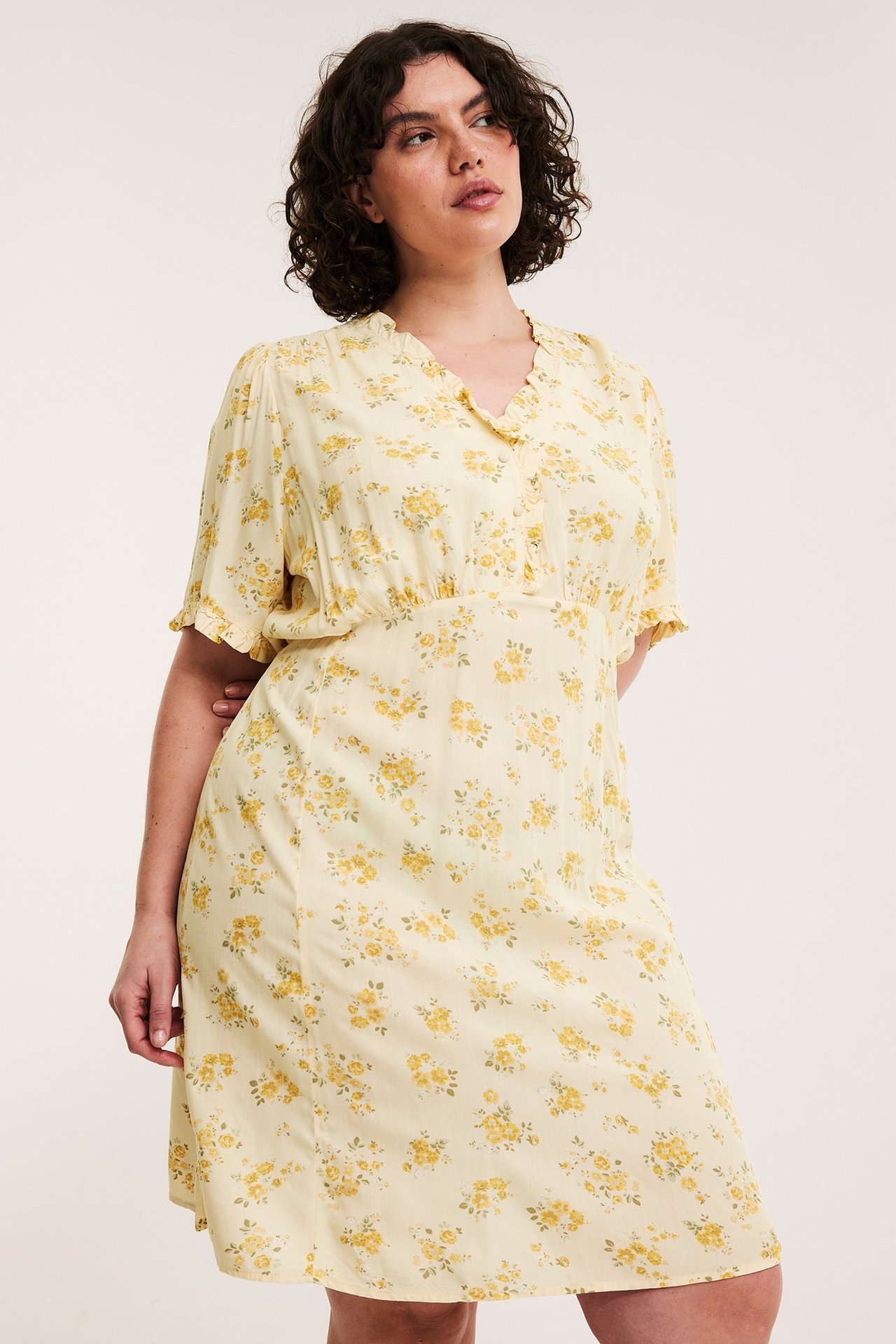 Edestä: nainen vaaleankeltaisessa kukkakuosisessa viskoosimekossa, polvipituinen, röyhelö V-pääntie ja lyhyet hihat.