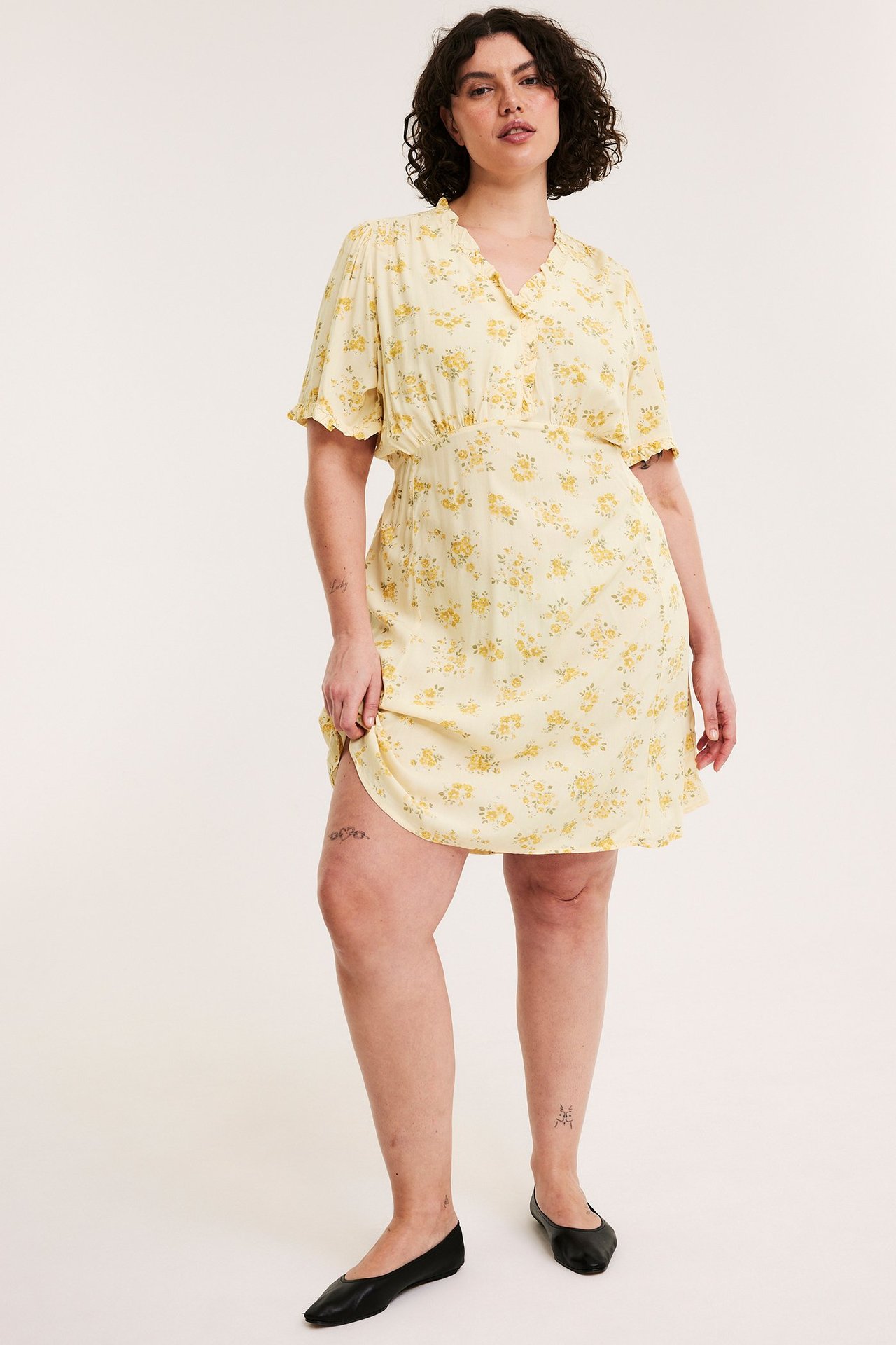 Edestäpäin: nainen vaaleankeltaisessa kukkakuosisessa viskoosimekossa, jossa röyhelöitä, ja mustat ballerinat.