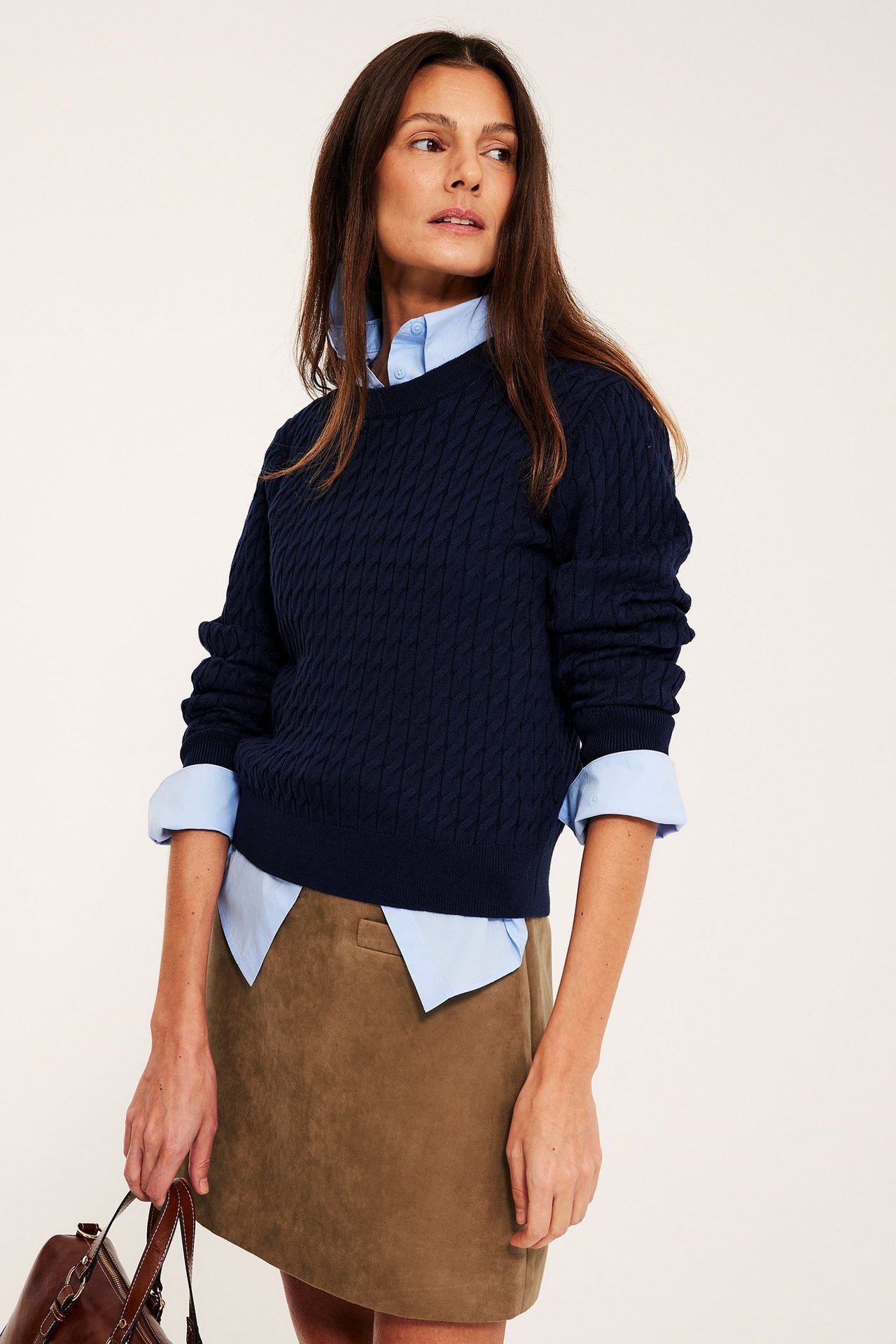 Nainen lyhyessä ruskeassa mokkajäljitelmähameessa, laivastonsinisessä neuleessa vaaleansinisen paidan päällä, pitäen ruskeaa laukkua.