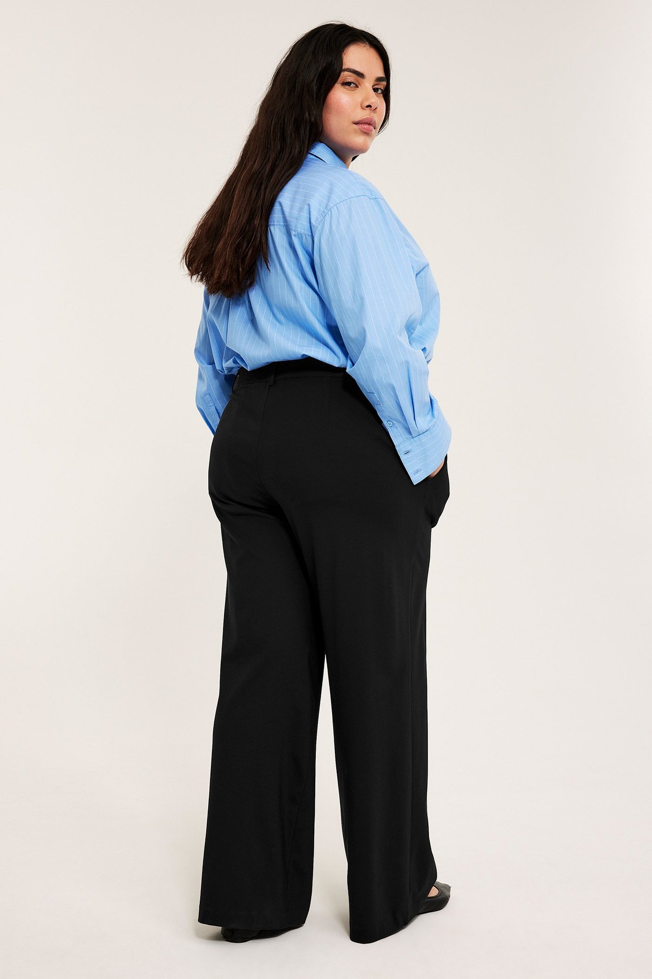 Kvinne sett bakfra i svarte vide bukser og blåstripet skjorte, ser over skulderen.