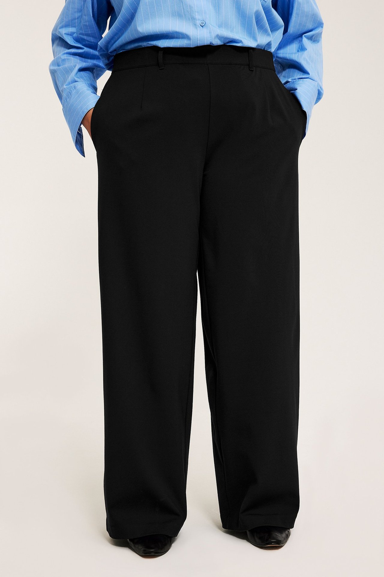 Framifrån: kvinna i svarta vida byxor, ljusblå randig skjorta och svarta platta skor.