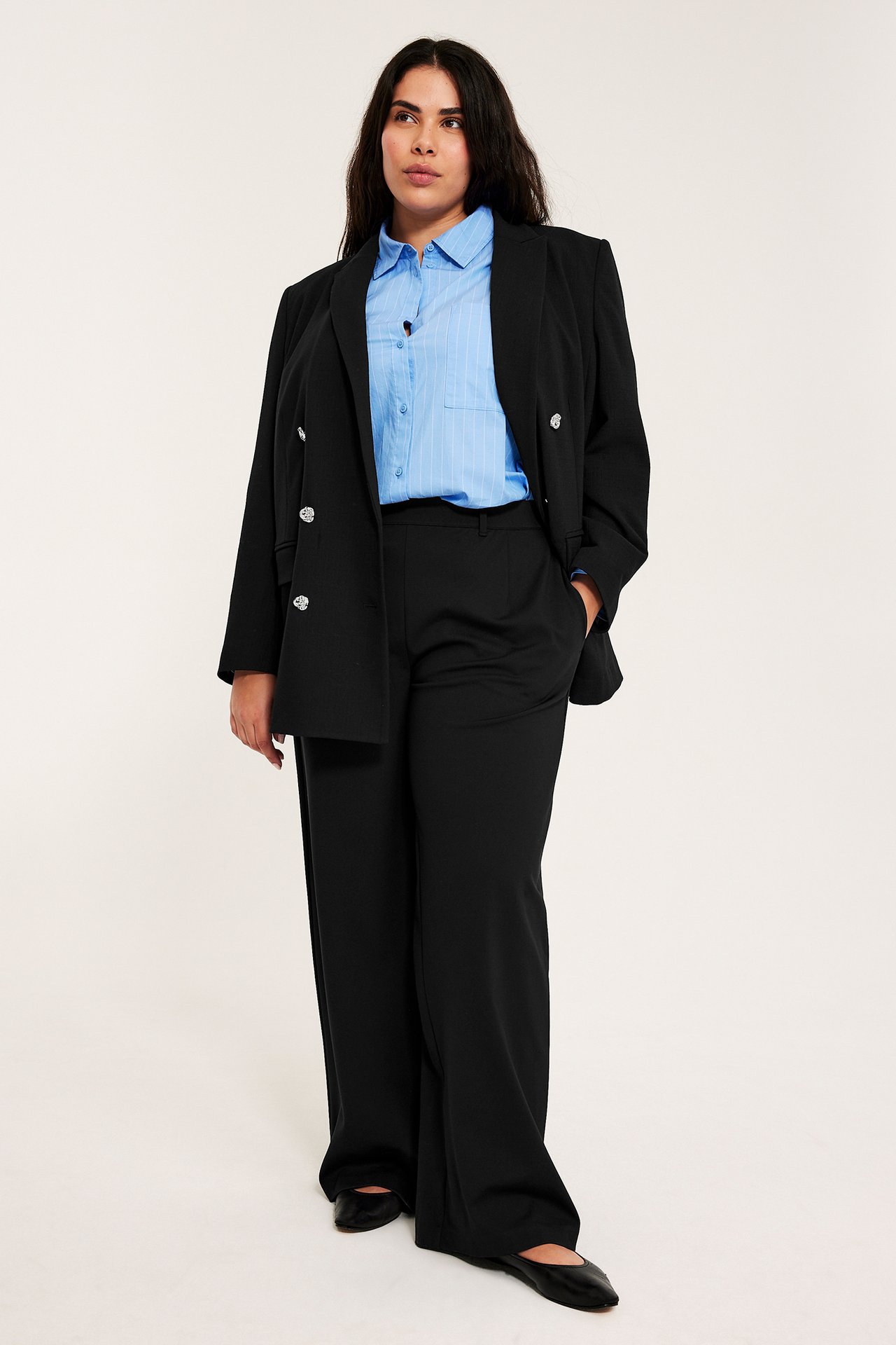 Etunäkymä: Nainen mustissa, leveälahkeisissa housuissa, vaaleansinisessä raitapaidassa ja mustassa bleiserissä.