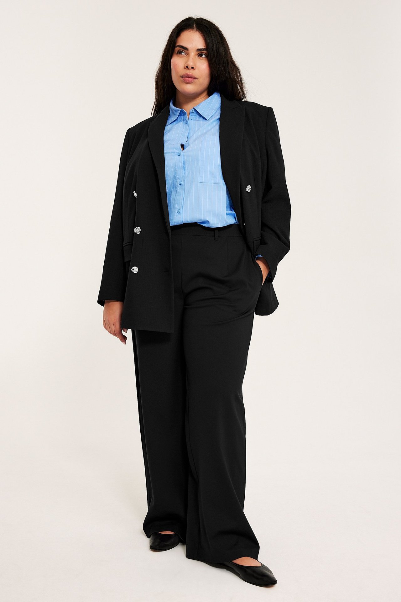 Frontvy: Kvinna i svarta vida dressbyxor, en ljusblå randig skjorta och en svart kavaj.