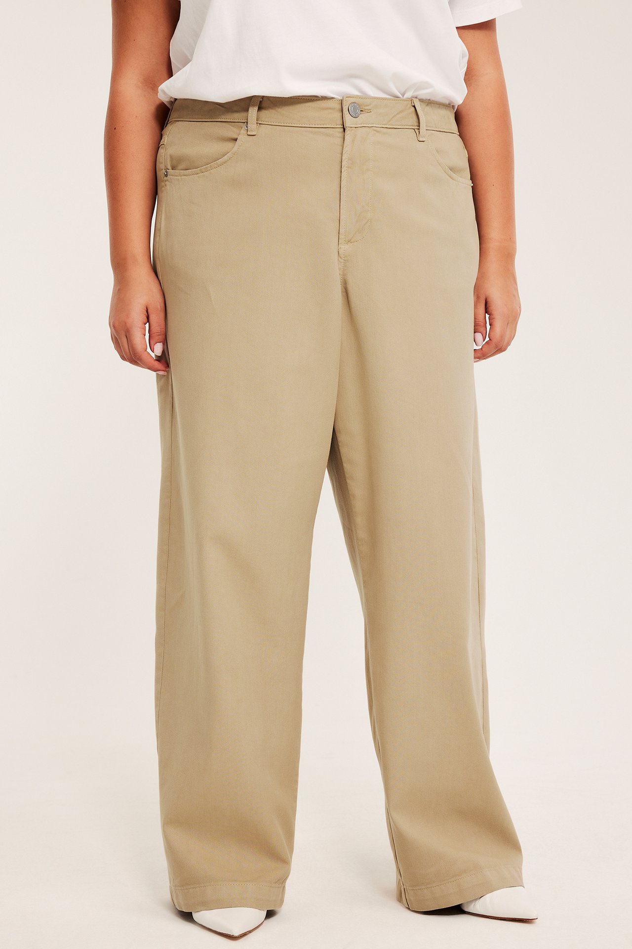 Frontbilde av en kvinne i beige vide bukser, stylet med en hvit t-skjorte og hvite spisse sko.