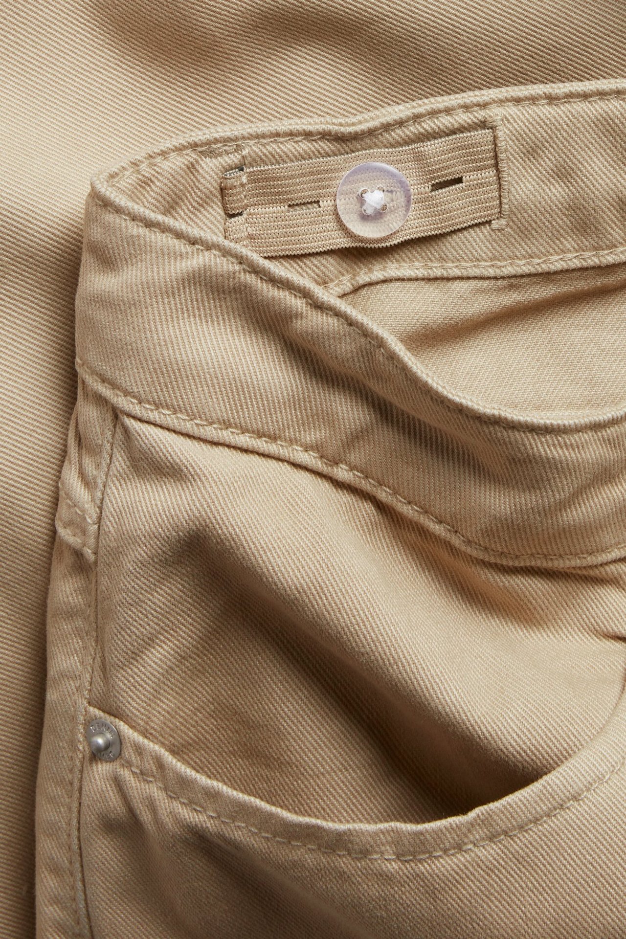 Zbliżenie beżowych szerokich spodni: pas z regulowaną gumką, zapięcie na guzik i przednia kieszeń.