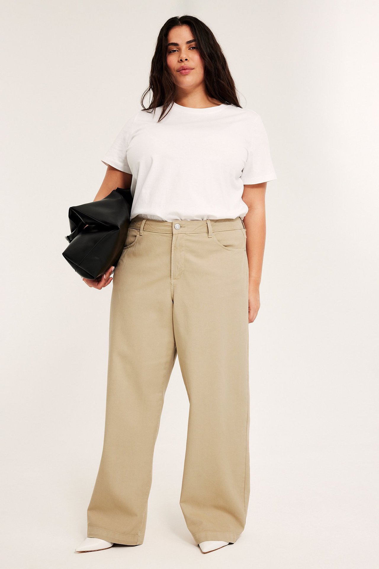 Frontvy av kvinna i vida beiga byxor, vit t-shirt, vita skor och håller en svart handväska.