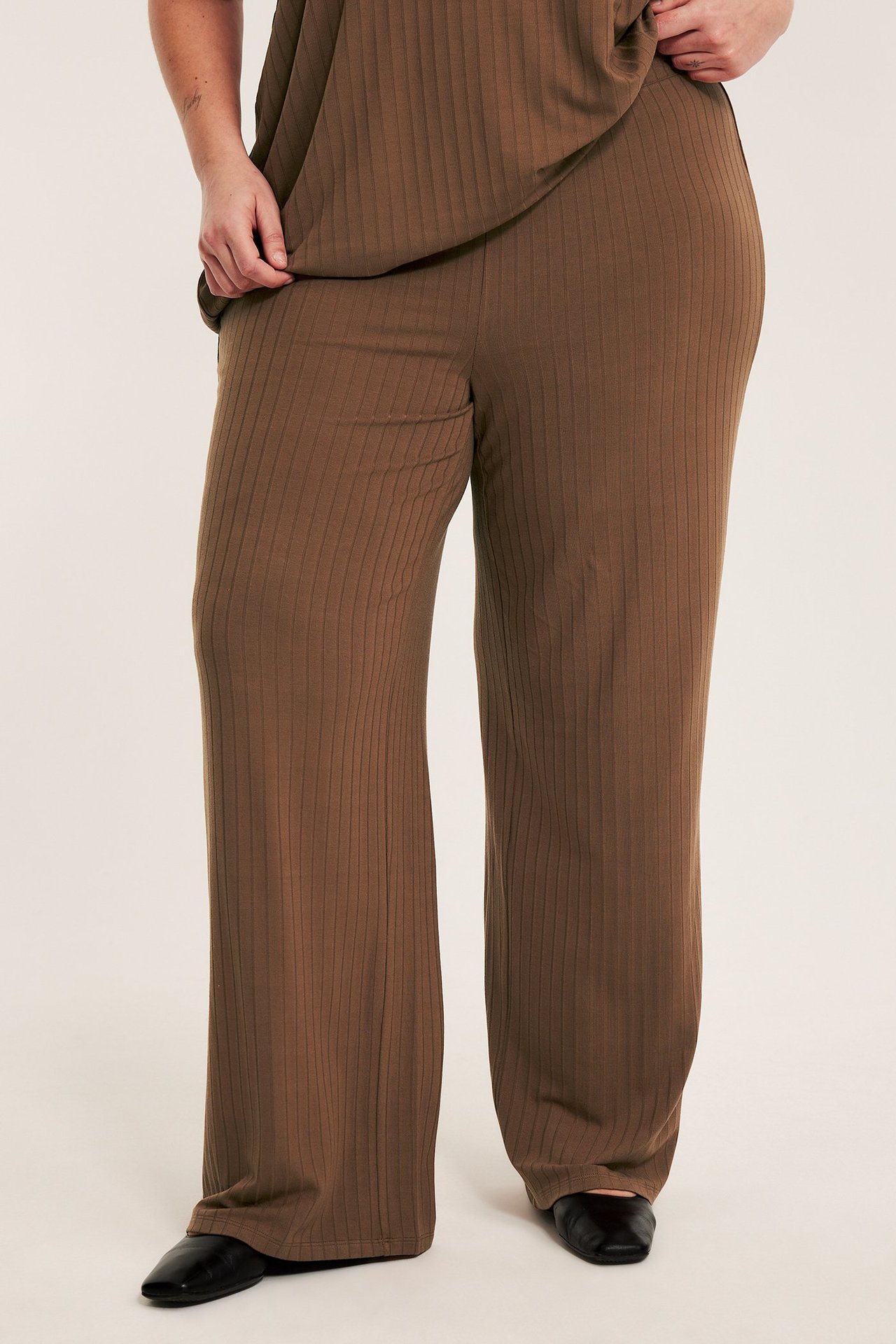 Widok z przodu: kobieta w brązowych prążkowanych szerokich spodniach, pasującym topie i czarnych butach.