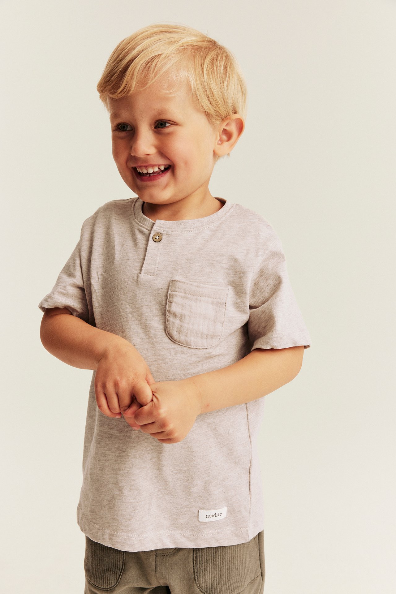 Framifrån: ett barn i ljusgrå kortärmad henley t-shirt med ficka och gröna manchesterbyxor.