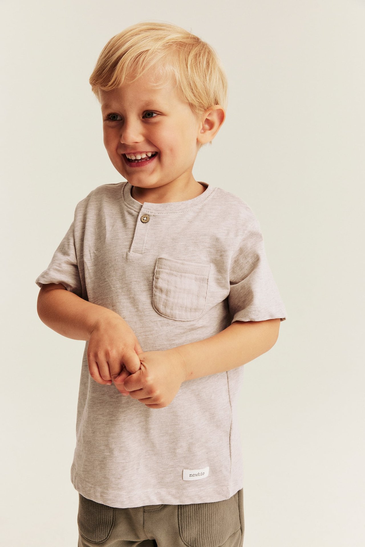 Framifrån: ett barn i ljusgrå kortärmad henley t-shirt med ficka och gröna manchesterbyxor.