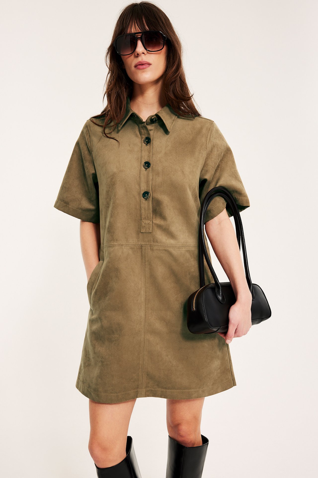 Kvinne bærer kort kjole i brunt imitert semsket skinn, holder svart veske, med svarte støvler og solbriller.