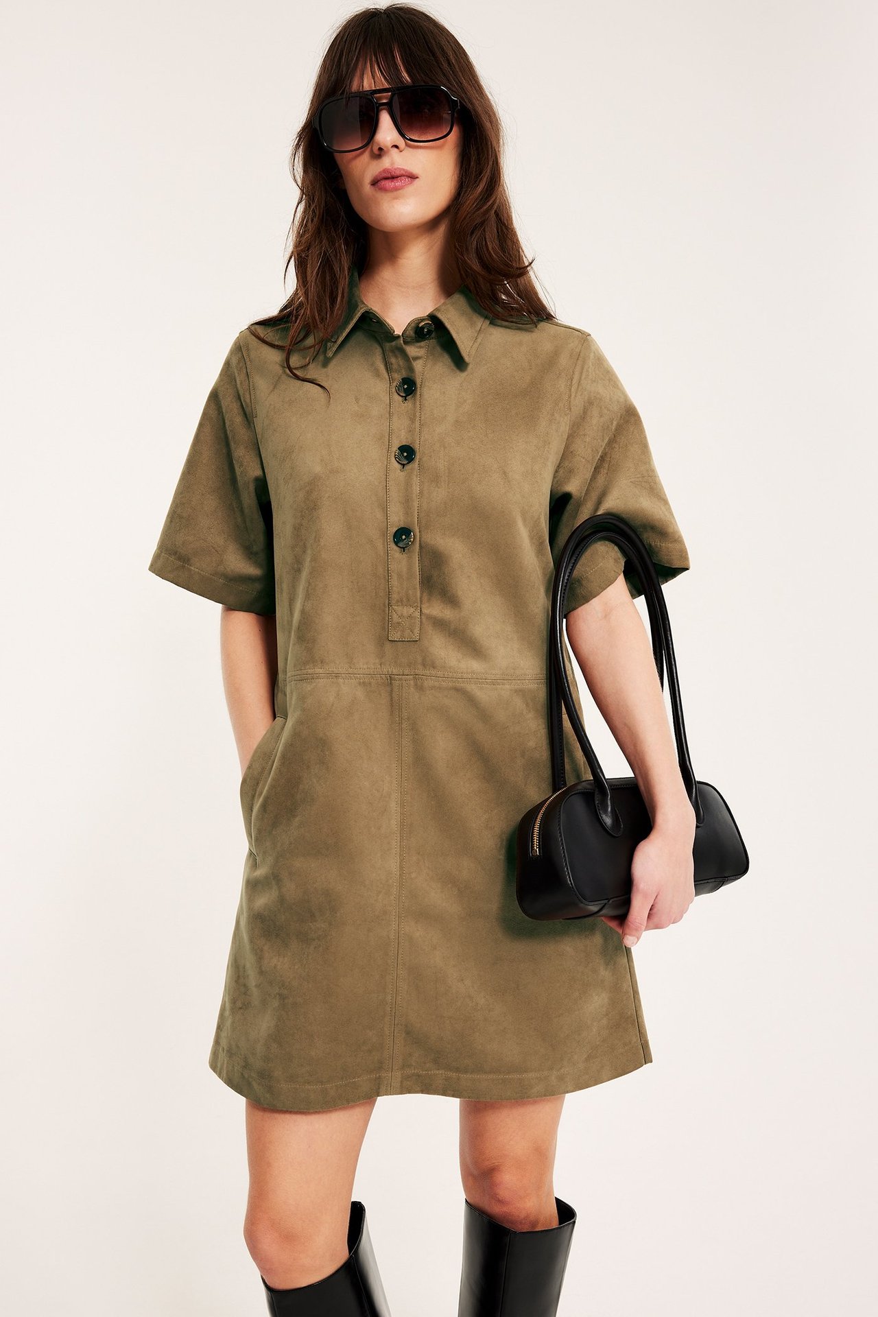 Kvinna bär kort klänning i brun mockaimitation, håller svart handväska, med svarta stövlar och solglasögon.