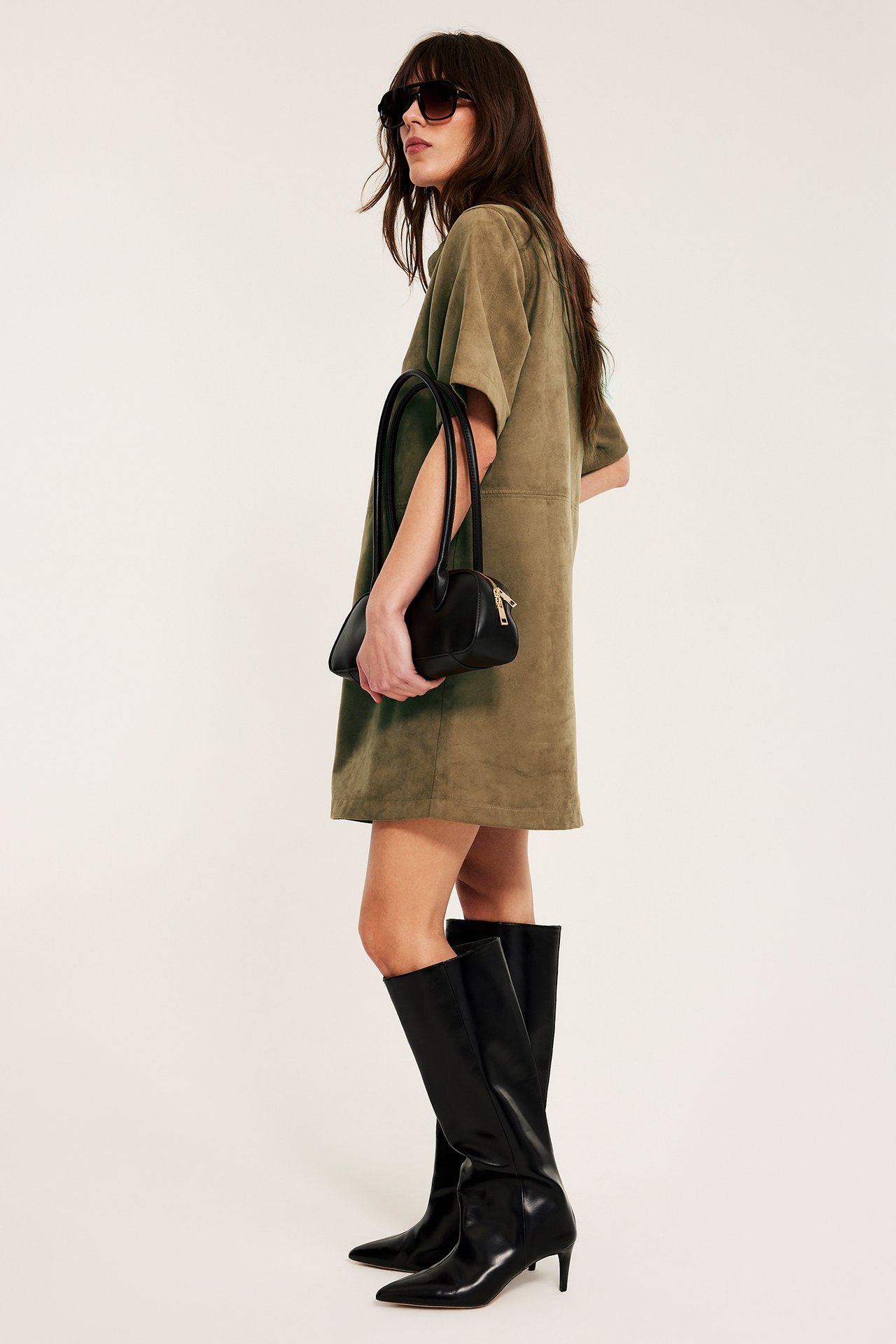 Sideutsikt: Kvinne i kort khaki-farget fuskemokkakjole, svarte knestøvler, med svart håndveske og solbriller.